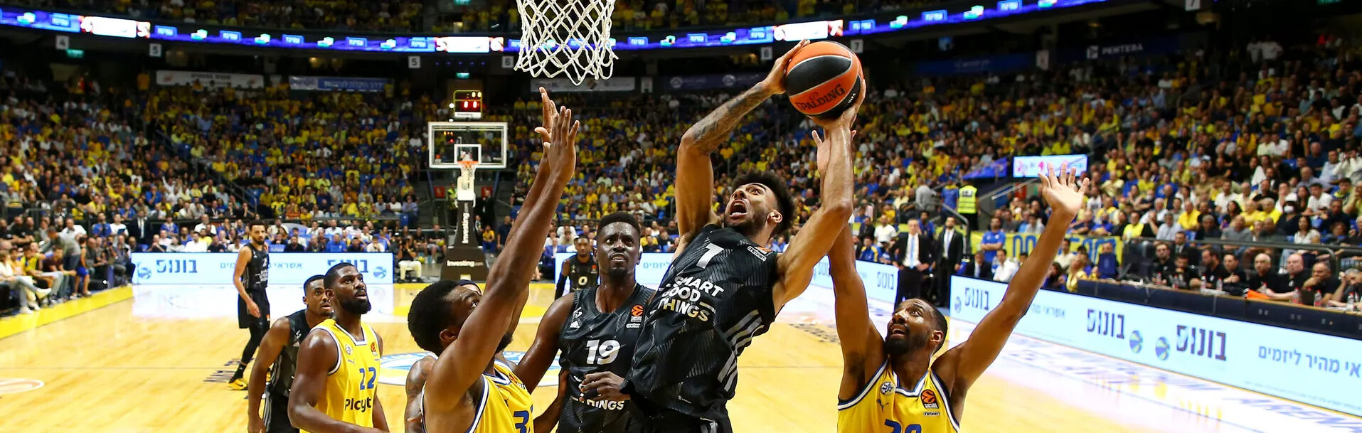 Basketballspieler in schwarzer Trikotfarbe springt zum Korb, um den Ball zu werfen, umgeben von mehreren Spielern in gelben Trikots in einer vollen Sportarena