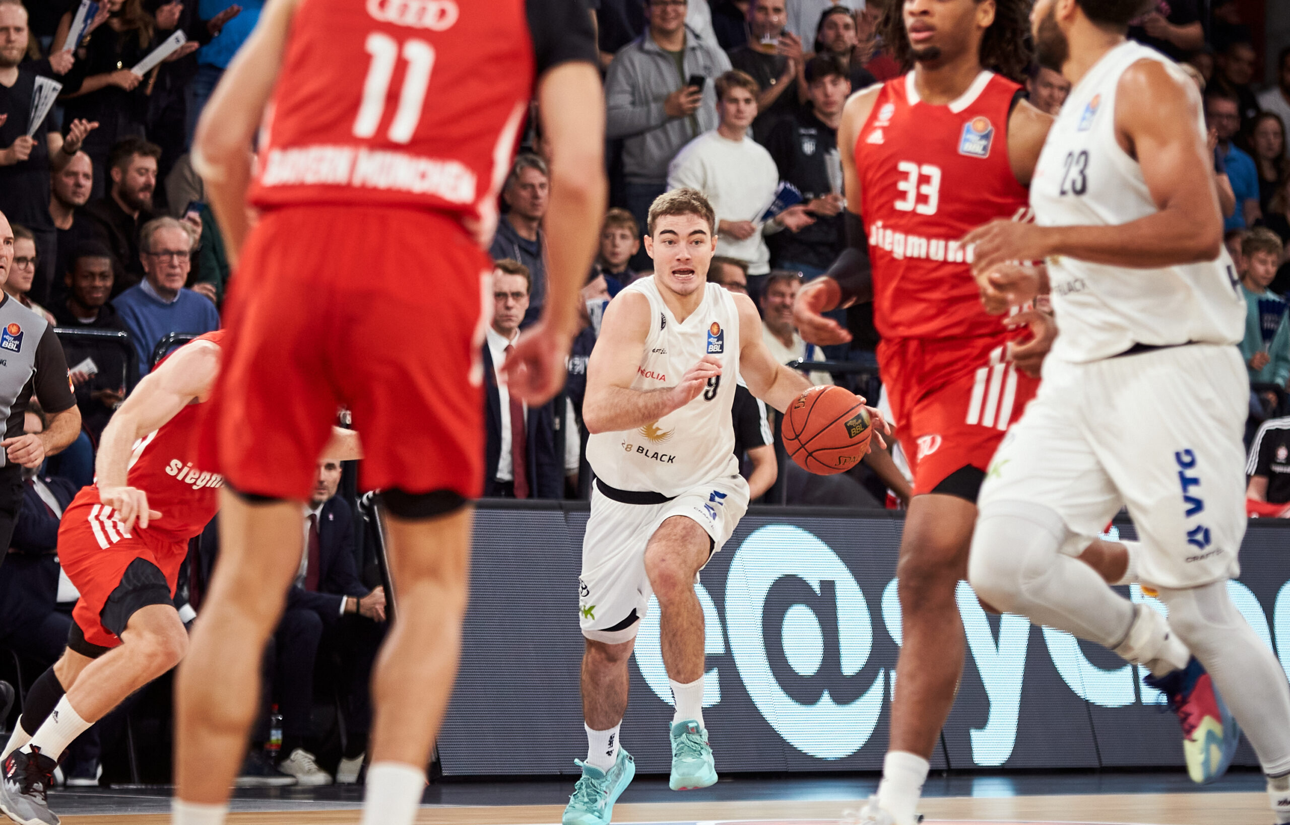 Basketballspieler in weißen Trikot mit Ball, umgeben von Spielern in roten Trikots während eines Spiels