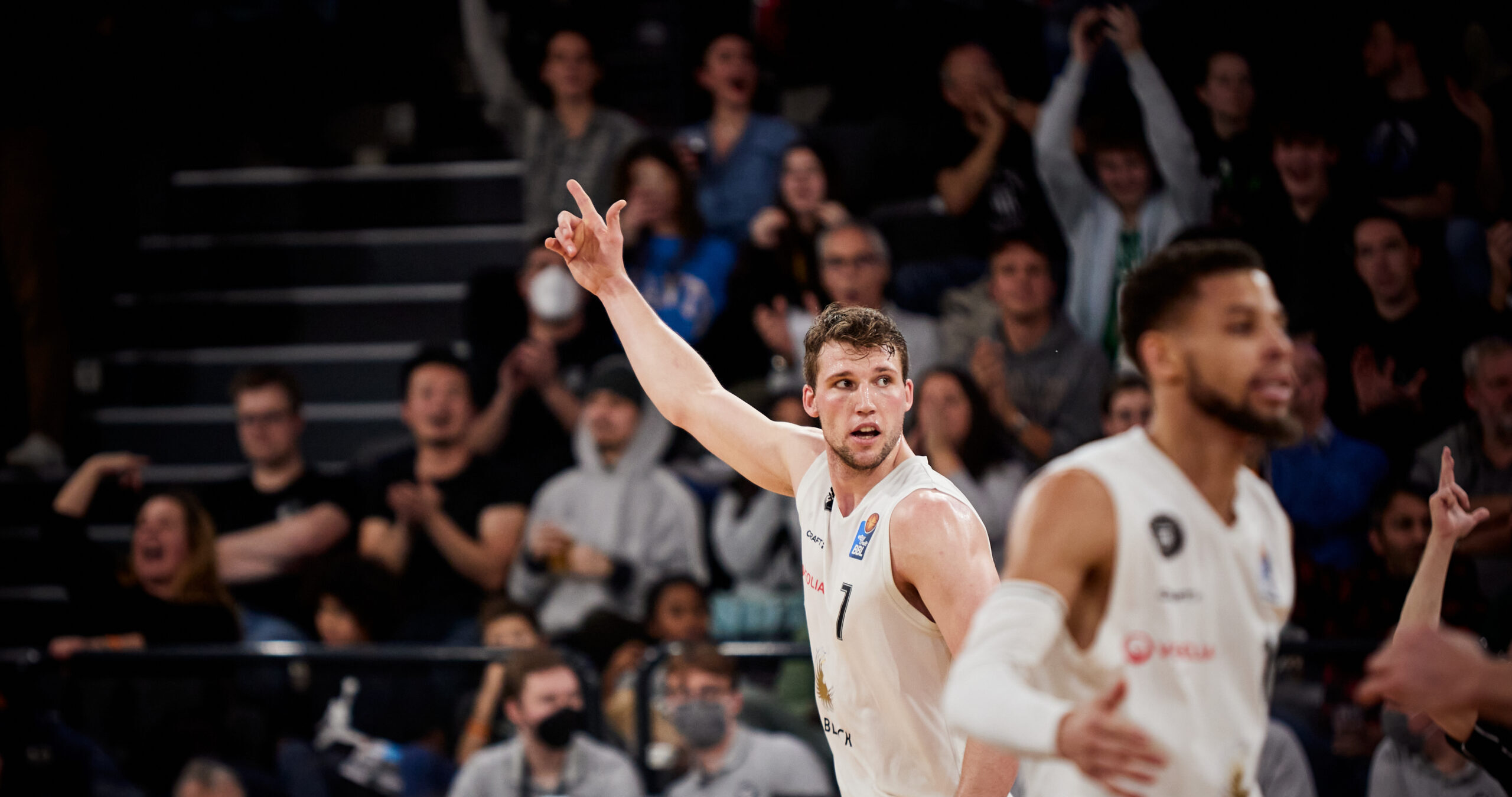 Basketballspieler mit Trikotnummer 7 hebt den rechten Arm und zeigt mit dem Zeigefinger nach oben während eines Spiels vor jubelndem Publikum