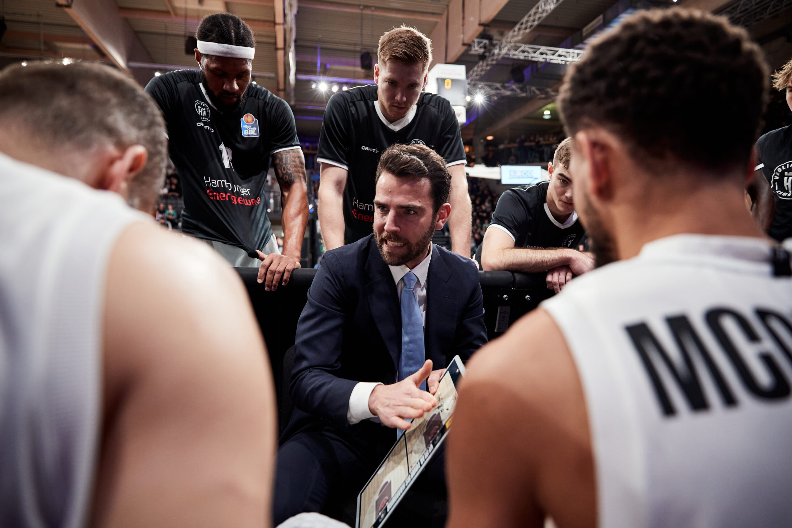 Basketballtrainer erklärt Taktik auf einem Whiteboard, umgeben von Spielern in weißen Trikots mit schwarzen Schriftzügen