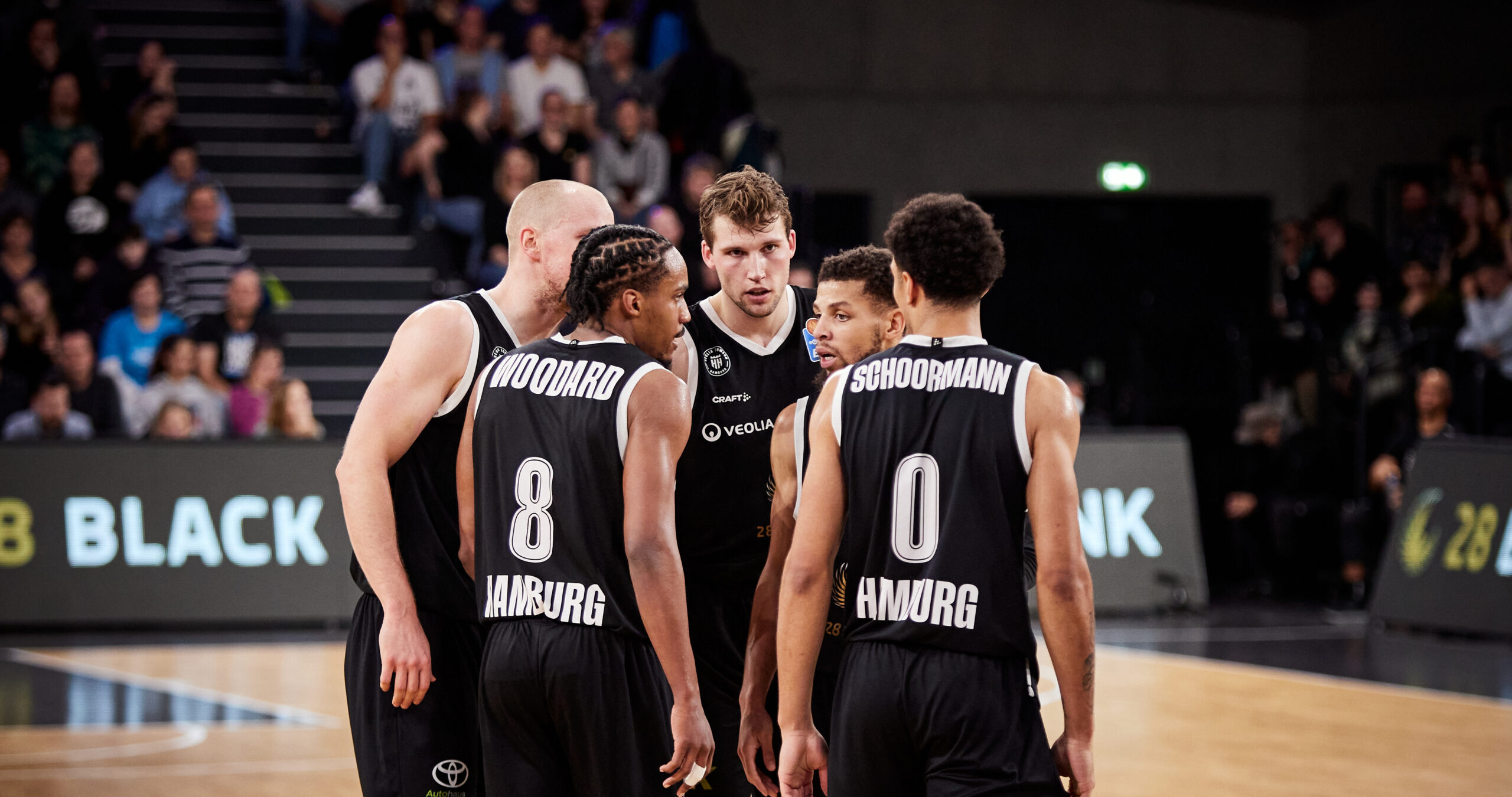 Basketballteam steht in einem Kreis zusammen, Spieler halten sich im Arm. Im Hintergrund eine Halle mit Fans