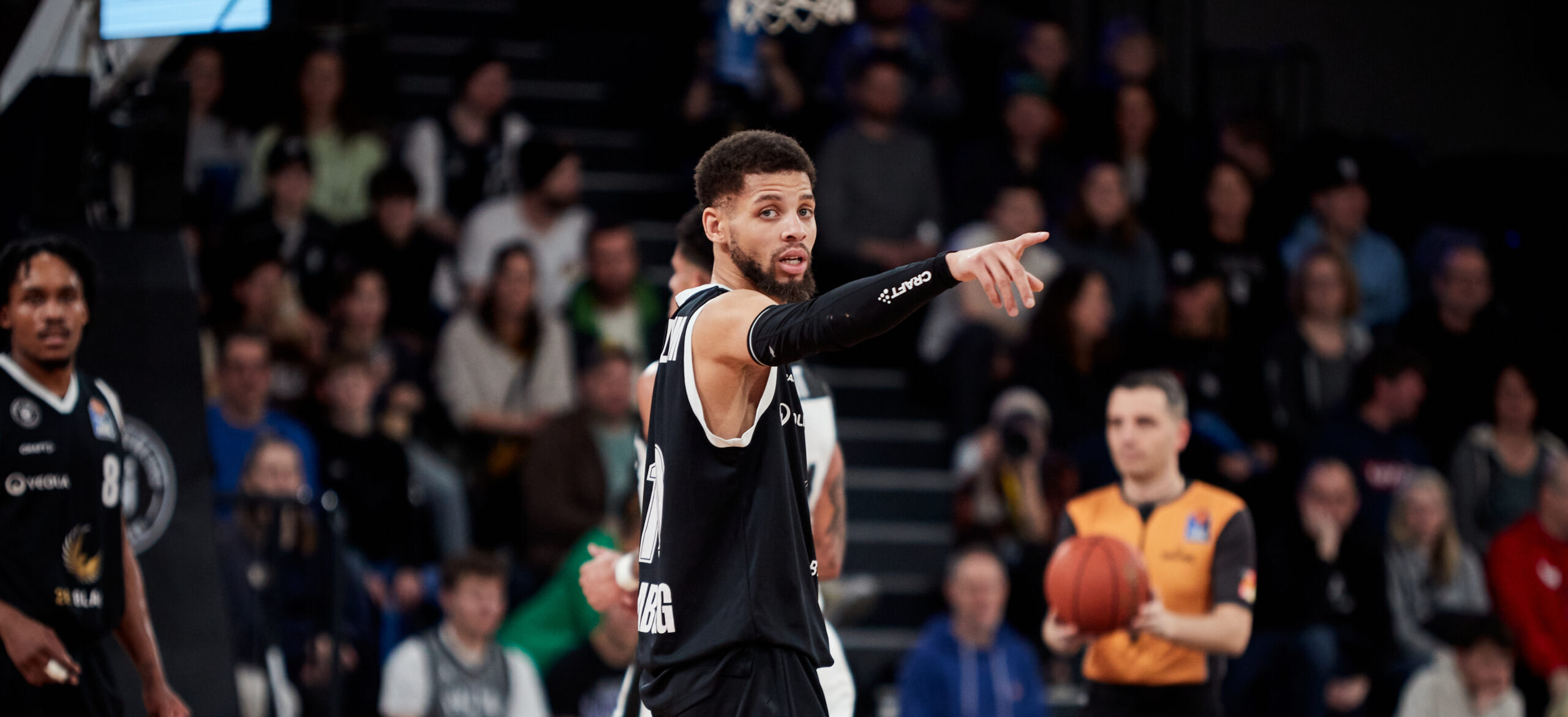 Basketballspieler in schwarzem Trikot mit Nummer 24 zeigt mit ausgestrecktem Arm während eines Spiels in einer Halle mit Zuschauern