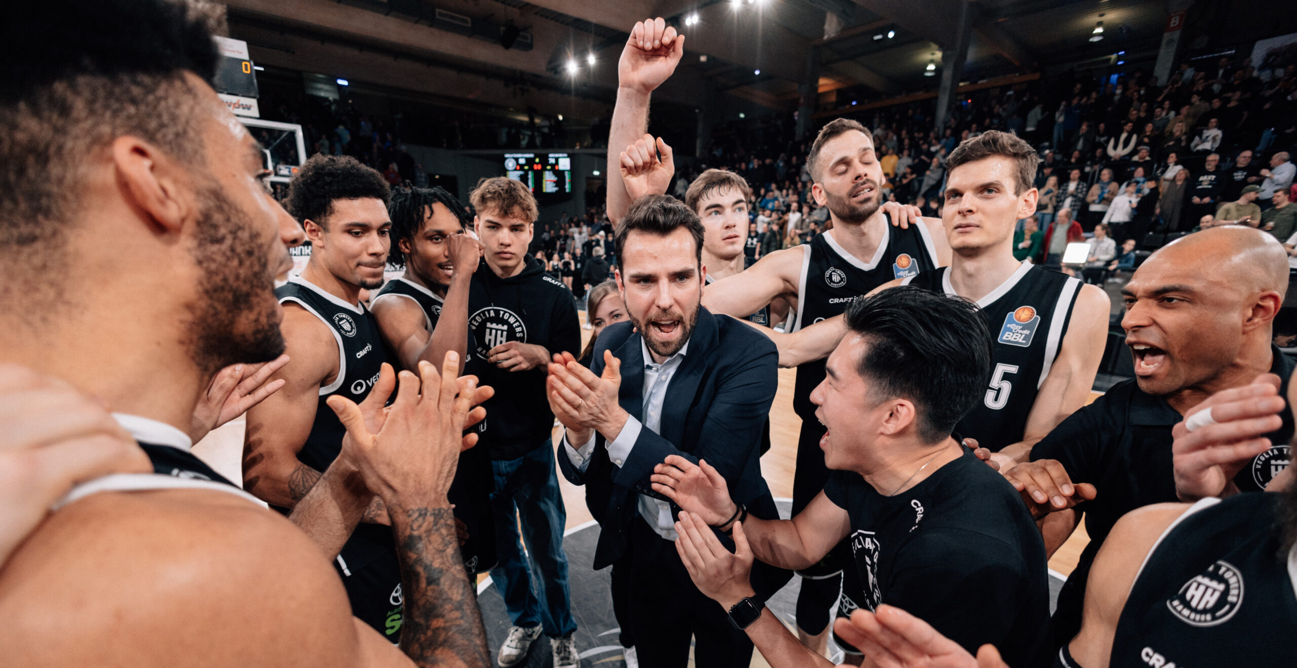 Basketballspieler und Trainer der Towers im Kreis, einige klatschen, einer hebt die Faust, Zuschauer im Hintergrund