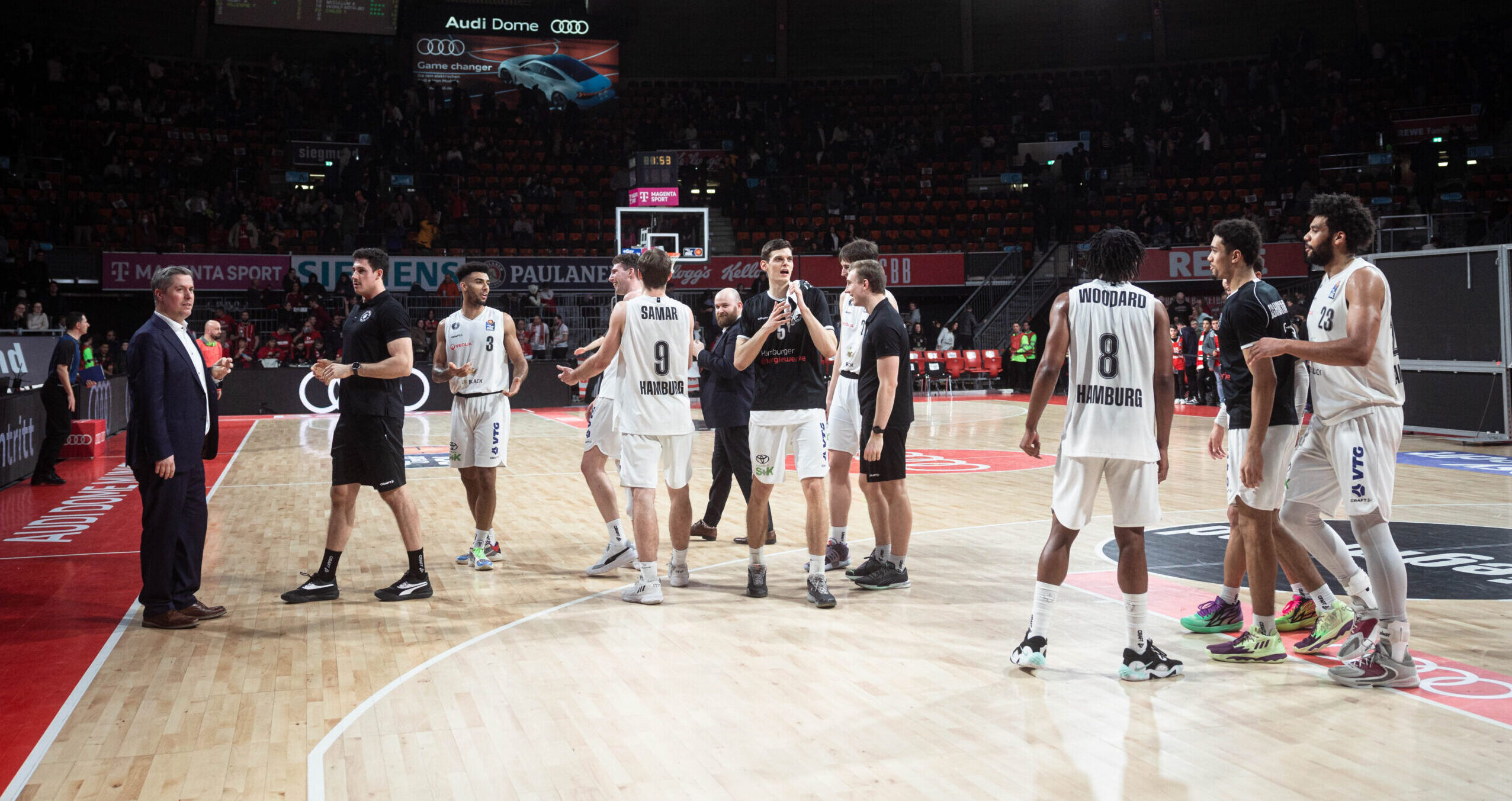 Spieler in weißen Trikots auf einem Basketballfeld klatschen in die Hände und umarmen sich, im Hintergrund dunkle Tribüne mit wenigen Zuschauern