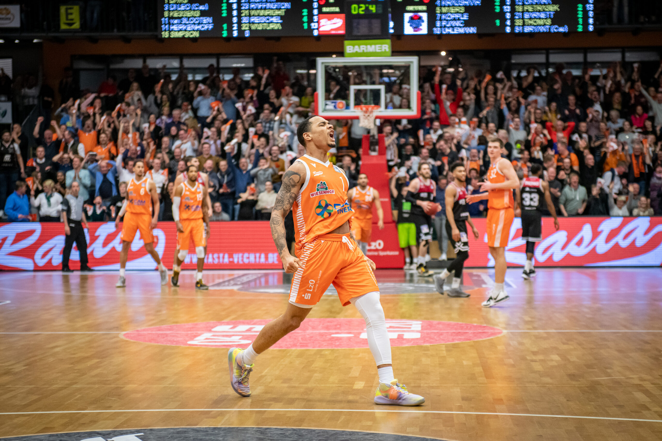 Basketballspieler in orangenem Trikot jubelt, im Hintergrund Fans und andere Basketballspieler