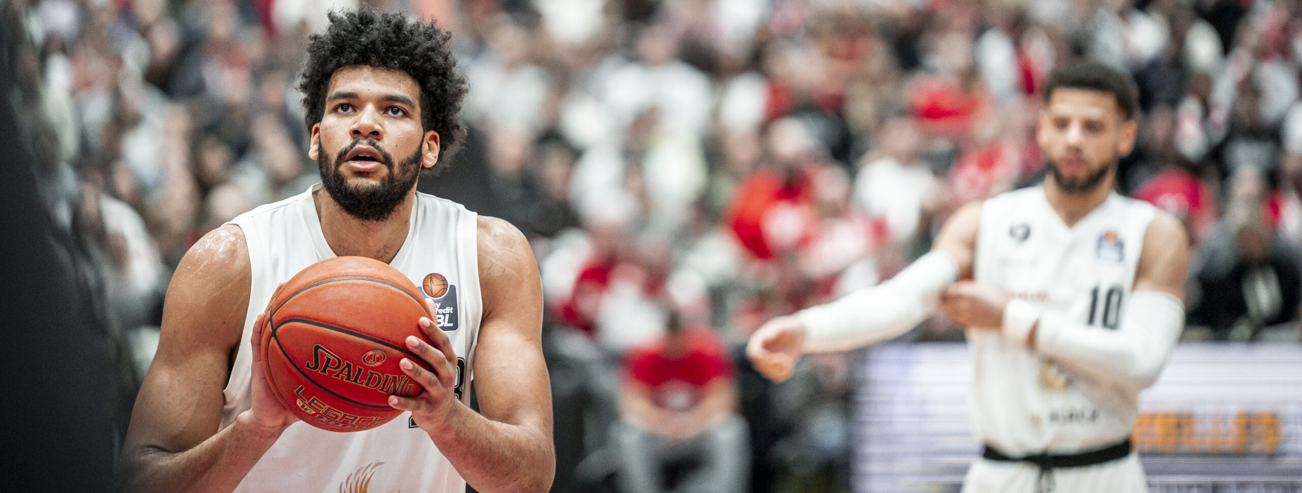 Basketballspieler in weißem Trikot hält einen Basketball vor sich, im Hintergrund ein weiterer Spieler und Zuschauer