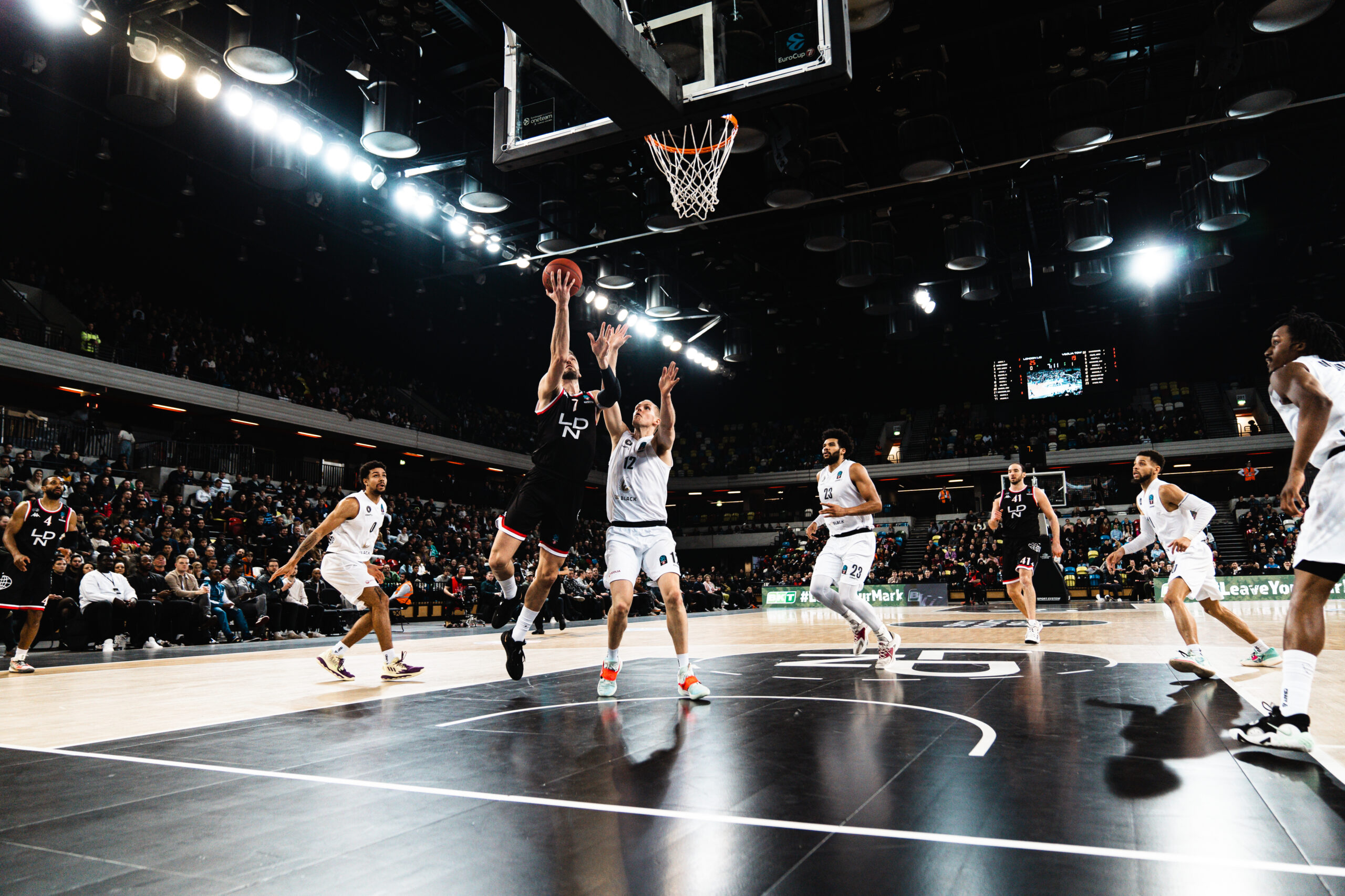 Basketballspieler in weiß und schwarz springen zum Korb, Zuschauertribüne im Hintergrund, Hallenbeleuchtung