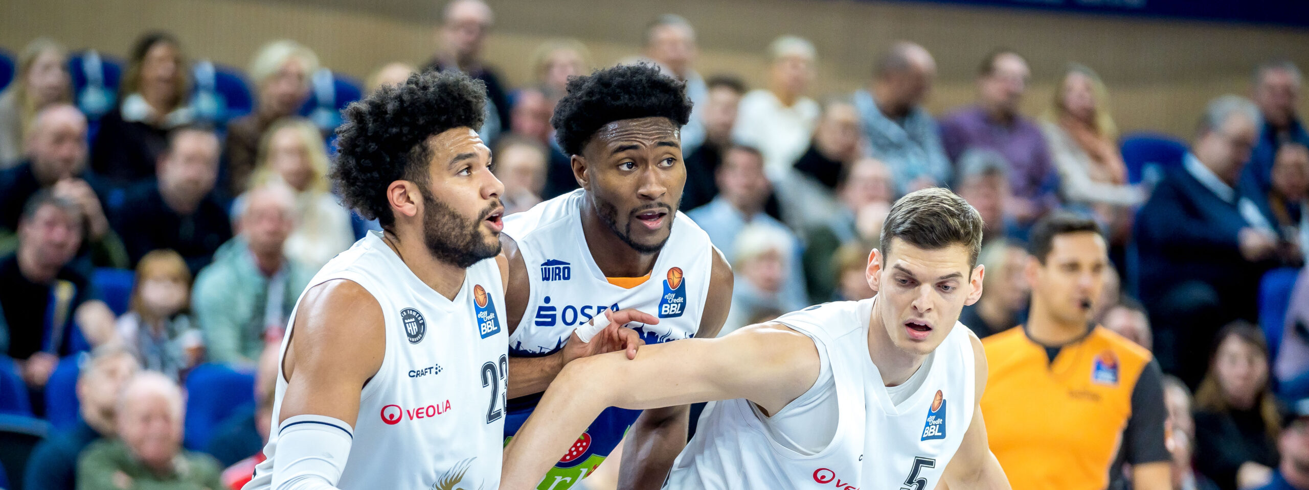 Zwei Basketballspieler in weißen Trikots mit Sponsorenlogos greifen sich an den Händen während eines Spiels