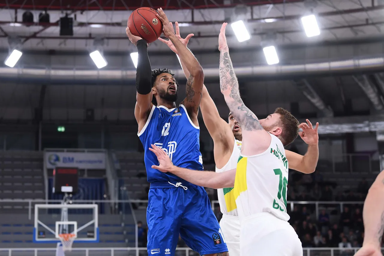 Basketballspieler in blauem Trikot springt zum Wurf, während zwei Spieler in weißen Trikots mit gelben und grünen Akzenten verteidigen