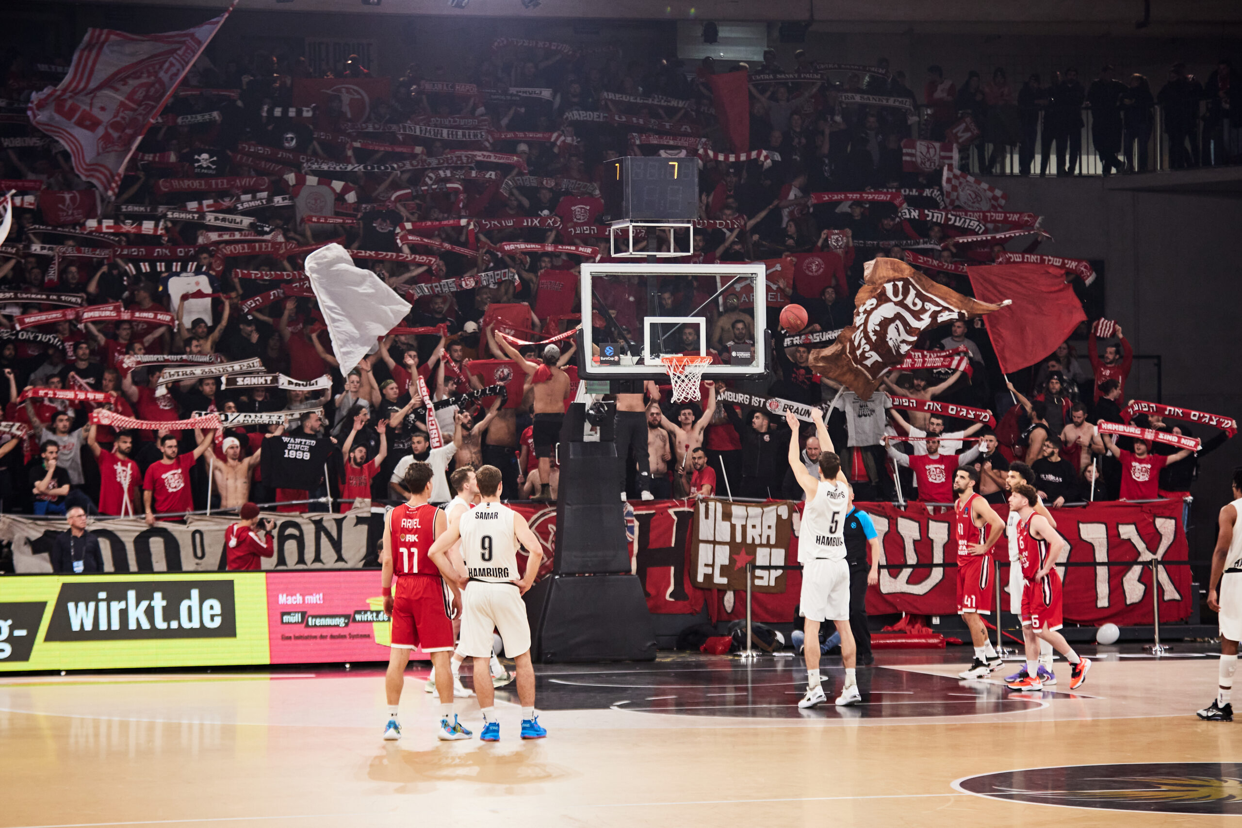 Basketballspieler in weißen Trikots beim Freiwurf vor Korb, umgeben von Spielern in roten Trikots, Fans mit Fahnen und Schals im Hintergrund