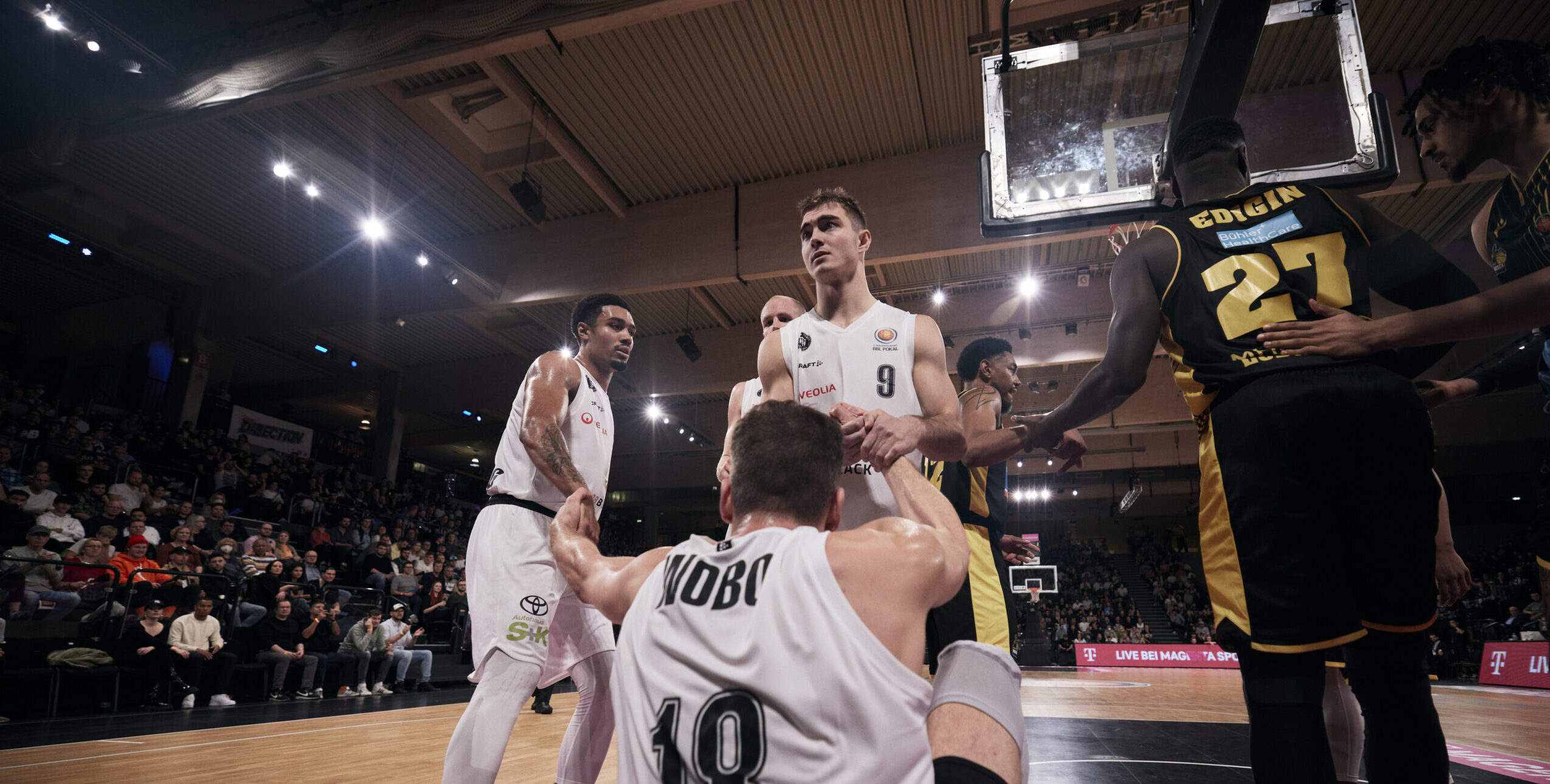 Basketballspieler mit Trikotnummer 10 sitzt auf dem Boden, während Teamkollegen und Gegner unter dem Korb stehen, Zuschauer im Hintergrund