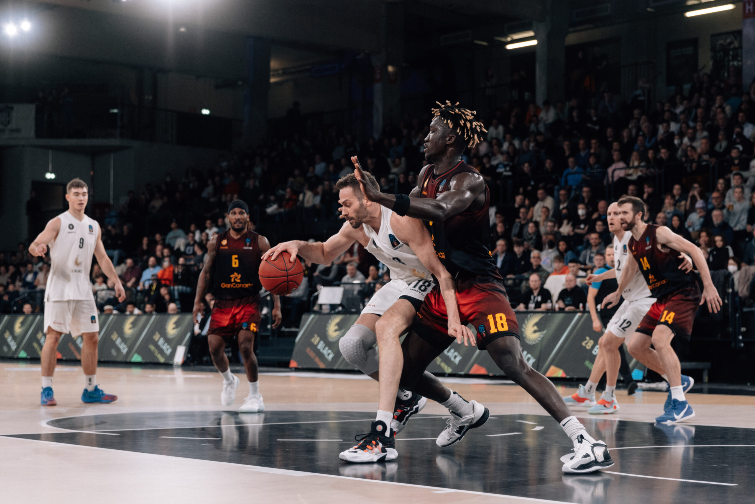 Basketballspiel zwischen zwei Teams, ein Spieler in weiß dribbelt den Ball, wird von einem Spieler in schwarzem Trikot mit der Nummer 18 verteidigt