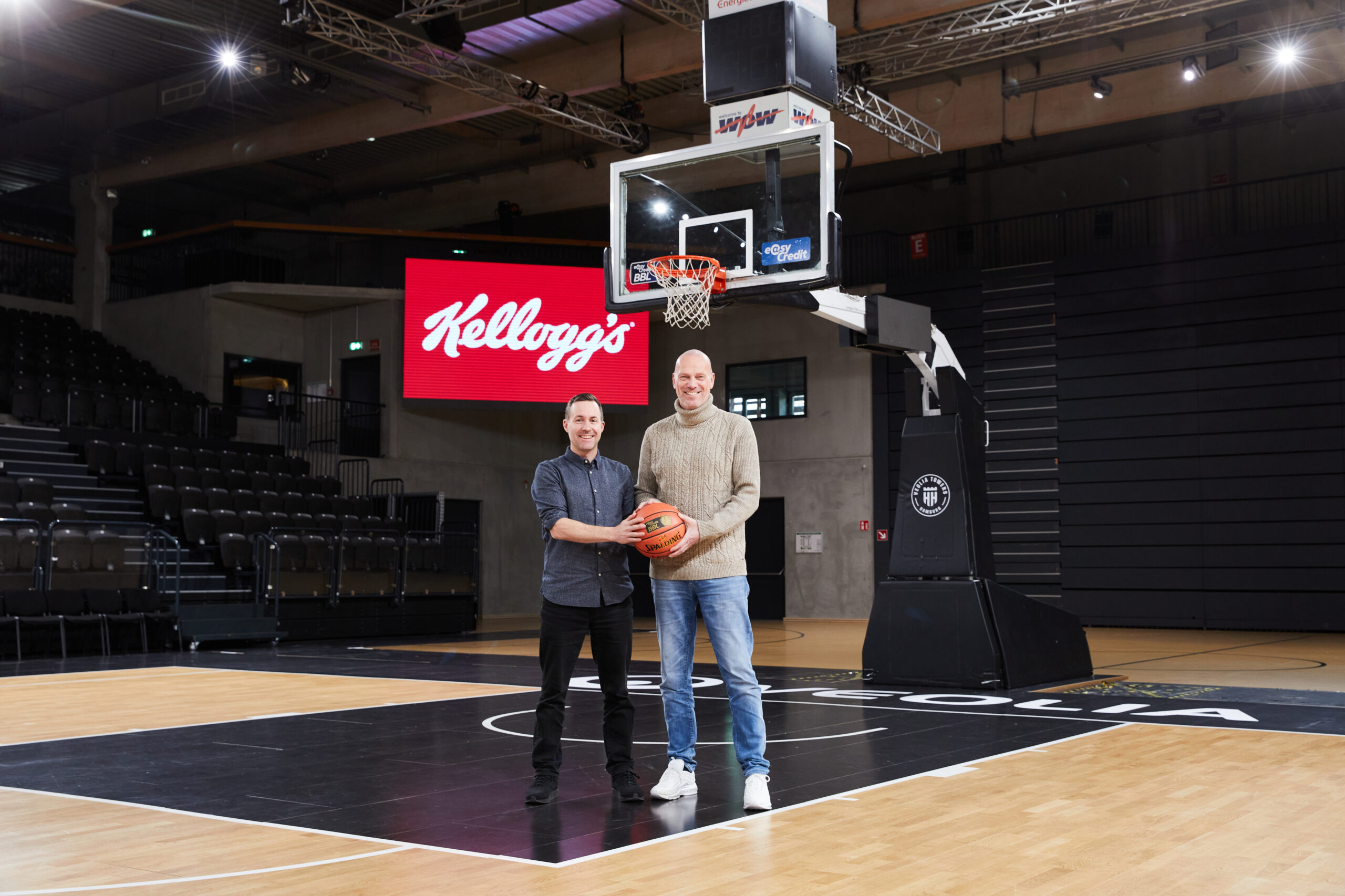 Zwei Männer stehen unter einem Basketballkorb, einer hält einen Basketball, im Hintergrund ein rotes Kellogg's-Logo