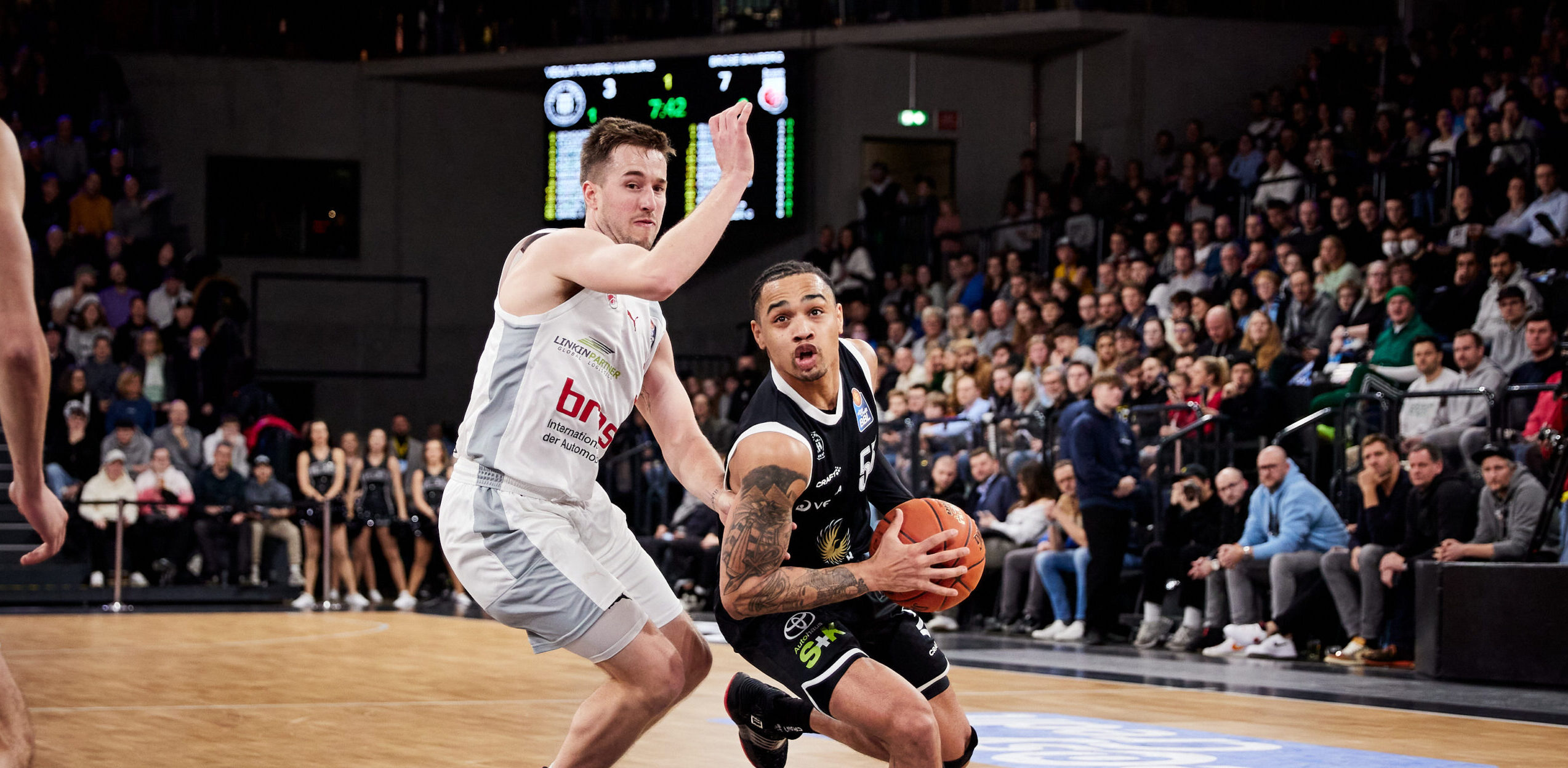 Basketballspieler in schwarzem Trikot mit Ball vor verteidigendem Spieler in weißem Trikot während eines Spiels vor voller Zuschauerkulisse