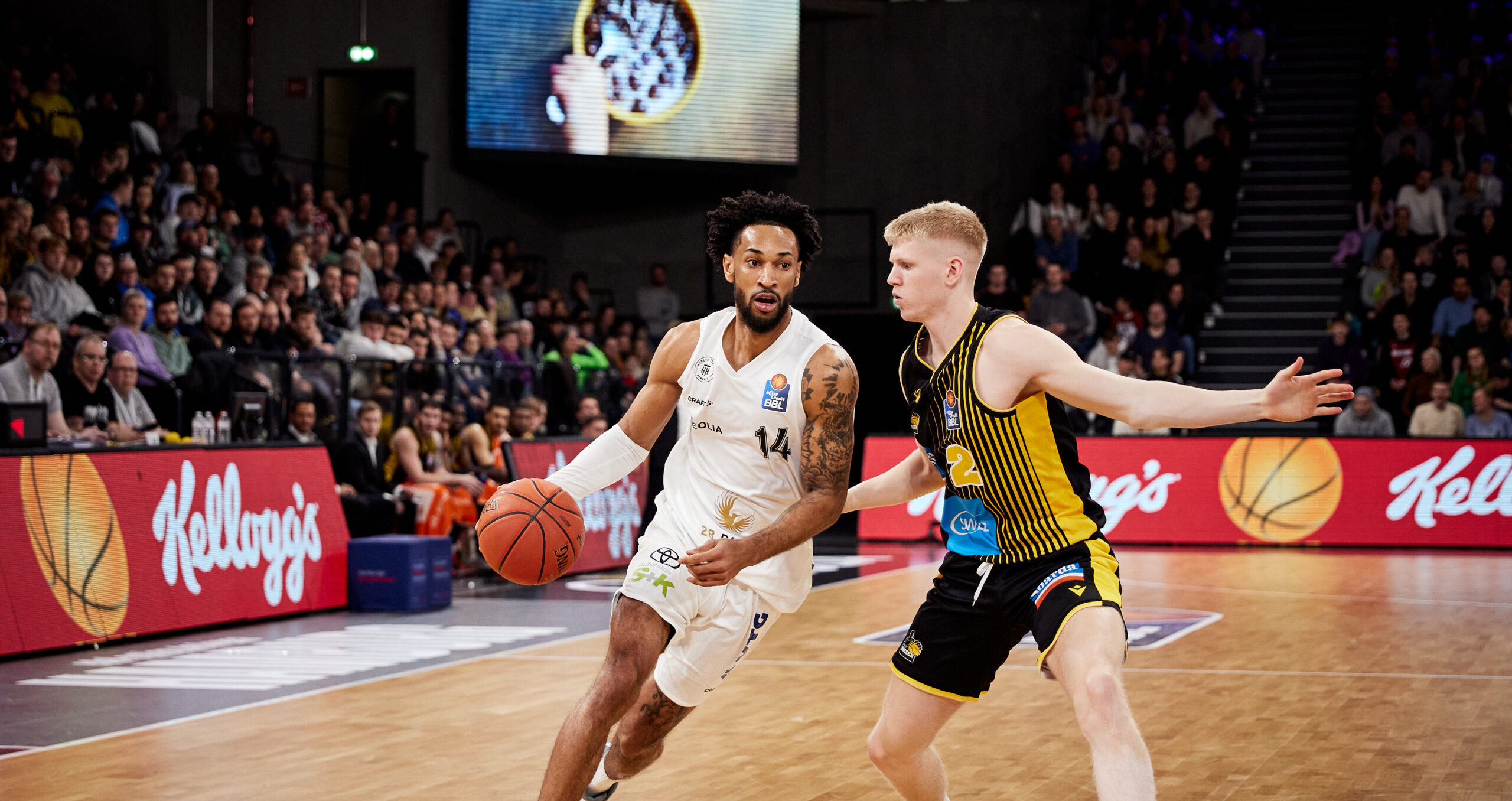 Basketballspieler in weißem Trikot mit Ball fährt am verteidigenden Spieler in schwarzem und gelbem Trikot vorbei während Zuschauer im Hintergrund zuschauen
