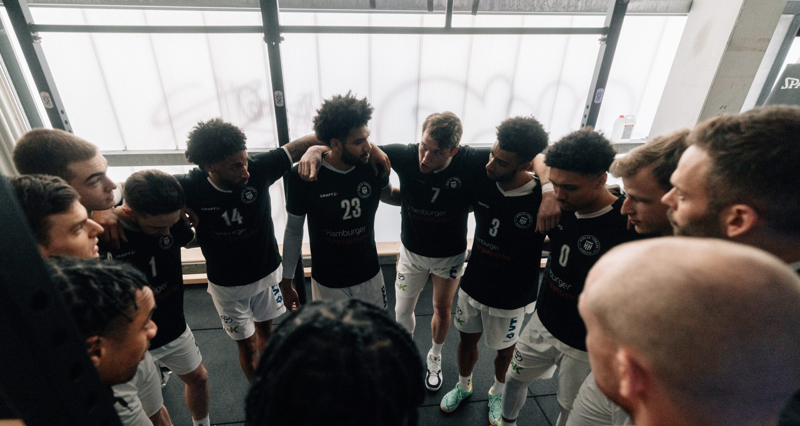 Basketballteam in schwarzen Trikots steht im Kreis, Arme auf Schultern gelegt, vor großen Fenstern in der Halle