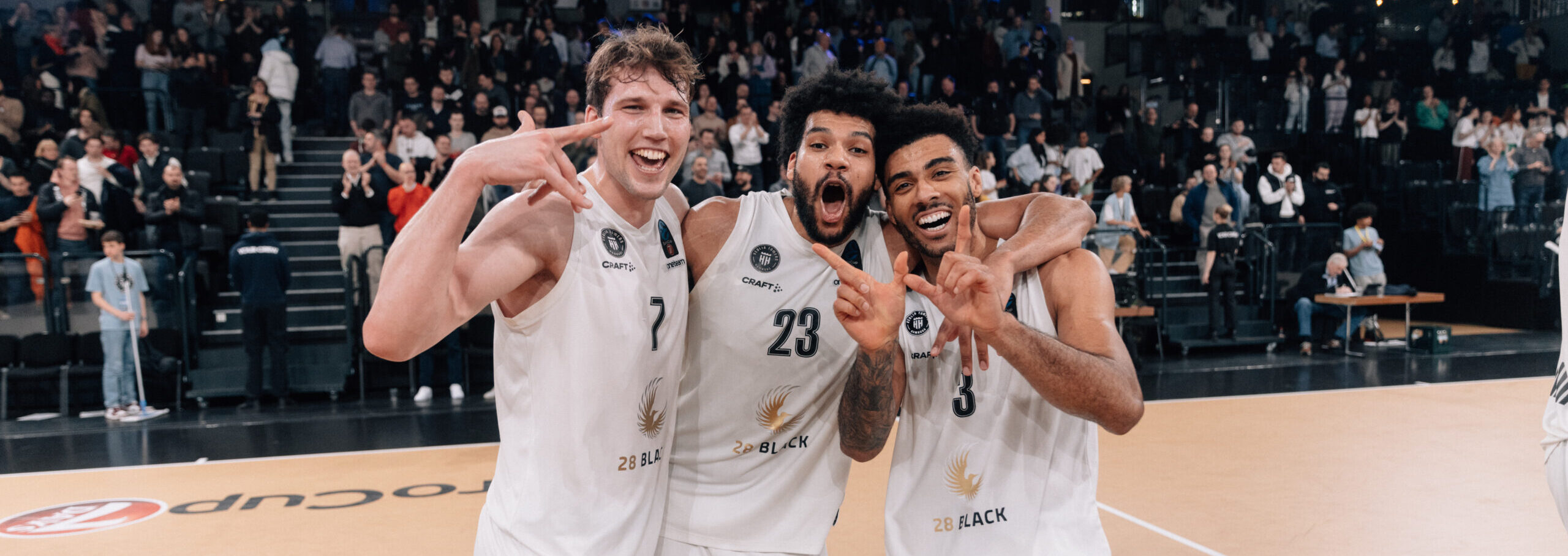 Drei Basketballspieler der Towers in weißen Trikots umarmen sich und zeigen Handzeichen auf dem Spielfeld vor jubelnden Zuschauern