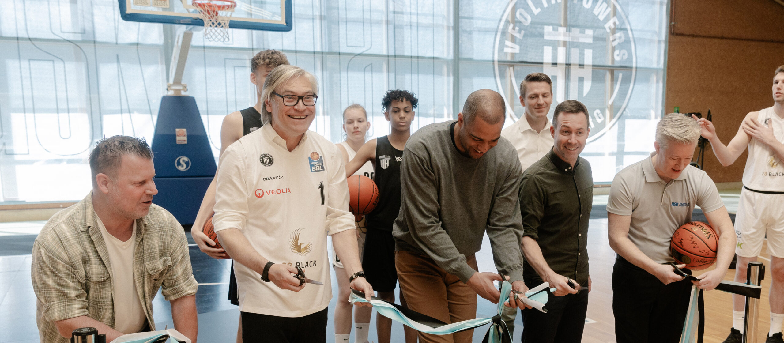 Mehrere Männer in einem Basketballtrainingszentrum, einer schneidet ein blaues Band durch, andere halten Basketbälle oder klatschen