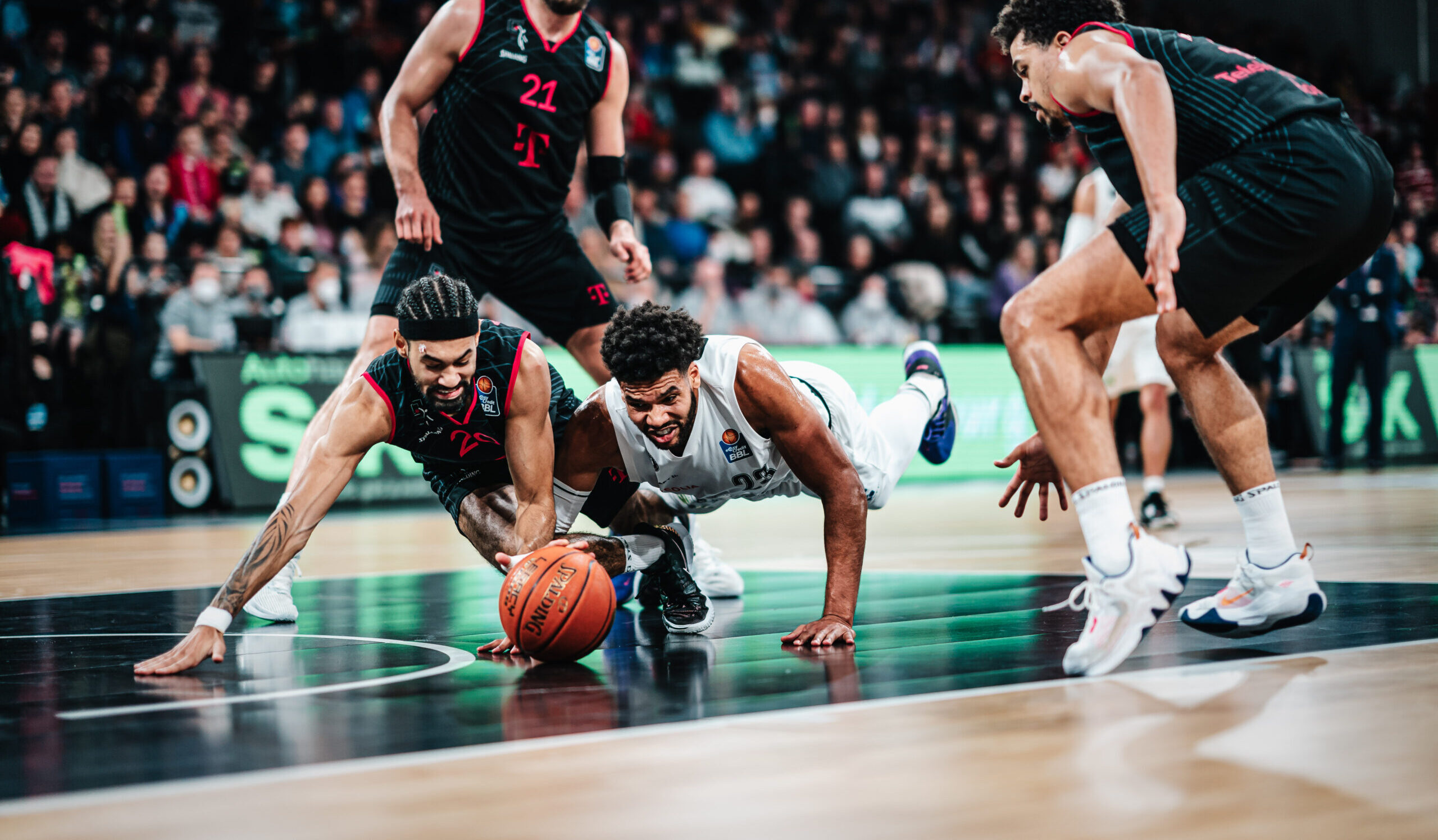 Spieler in weißen und schwarzen Trikots kämpfen auf dem Hallenboden um einen Basketball, weitere Spieler in schwarzen Trikots sowie Halle mit Zuschauern im Hintergrund