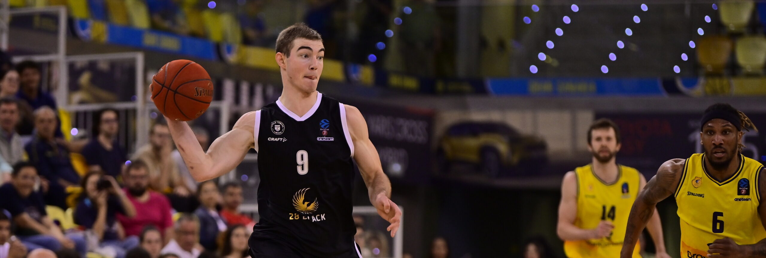 Basketballspieler mit Trikot Nummer 9 in schwarzer Uniform hält den Ball während eines Spiels in einer Halle