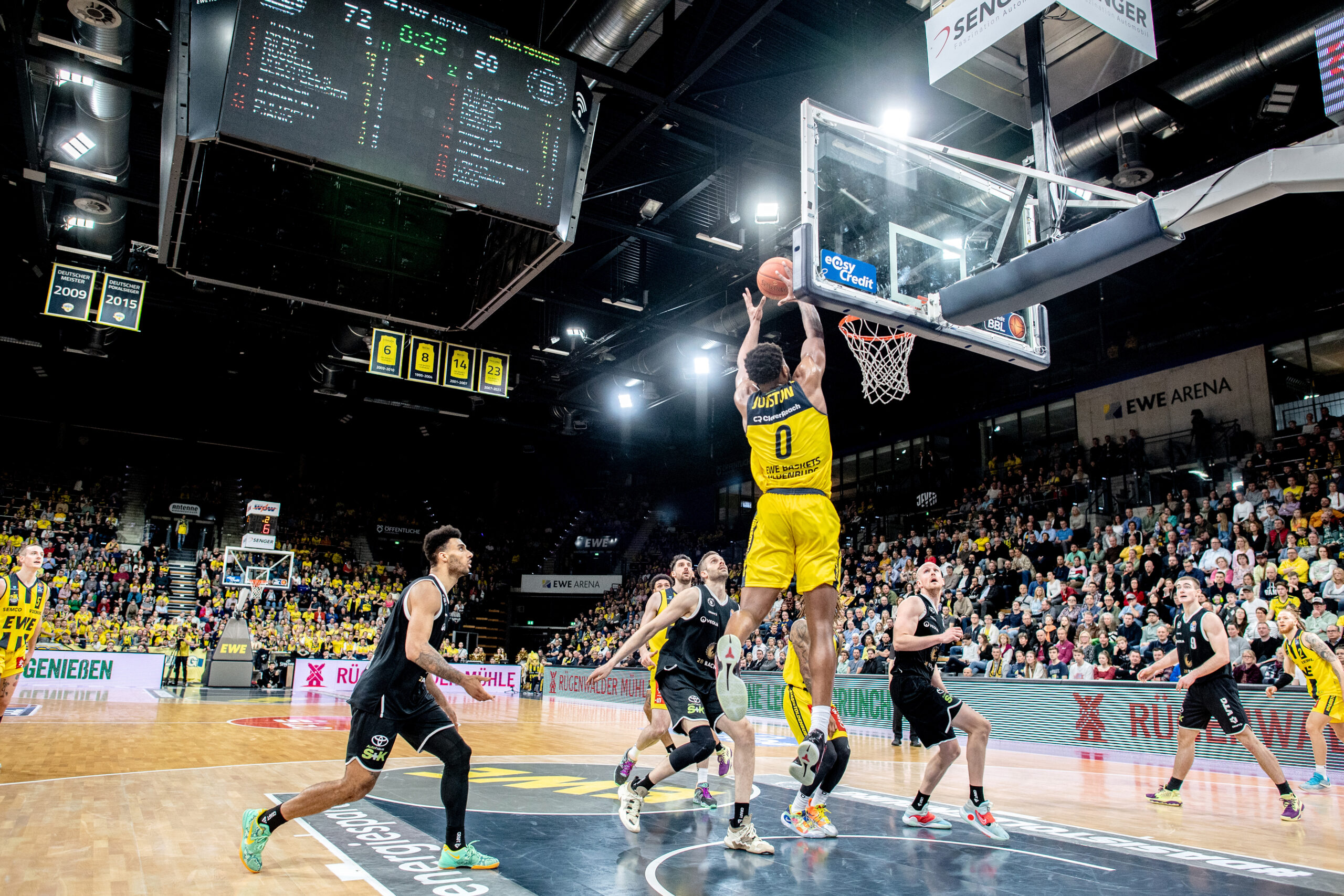 Spieler in gelbem Trikot springt mit Ball hoch zum Korb. Um ihn herum andere Spieler in schwarzen und gelben Trikots. Im Hintergrund eine Halle, an der Decke eine Anzeigetafel
