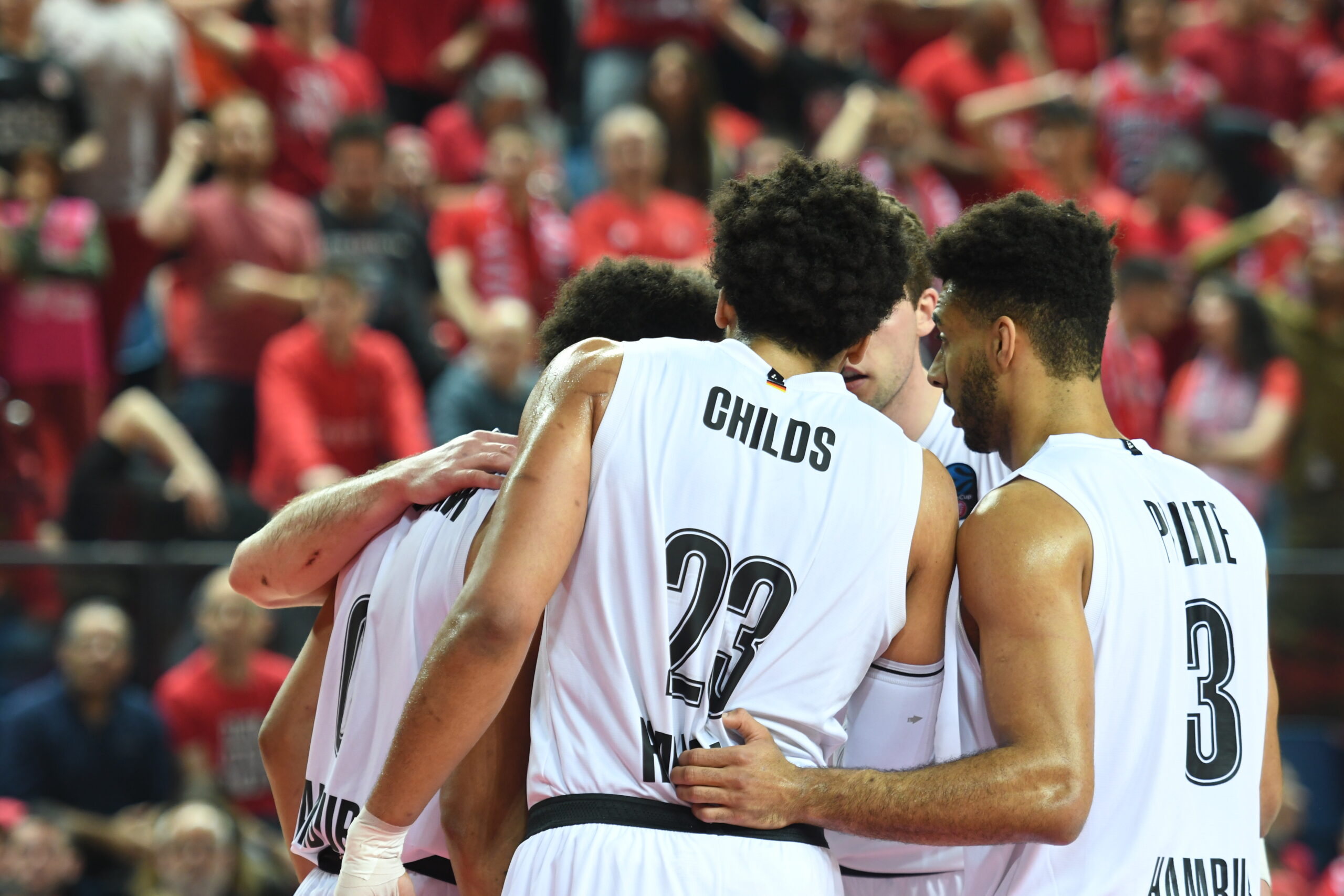 Vier Basketballspieler in weißen Trikots umarmen sich während eines Spiels vor Zuschauern in roten Shirts