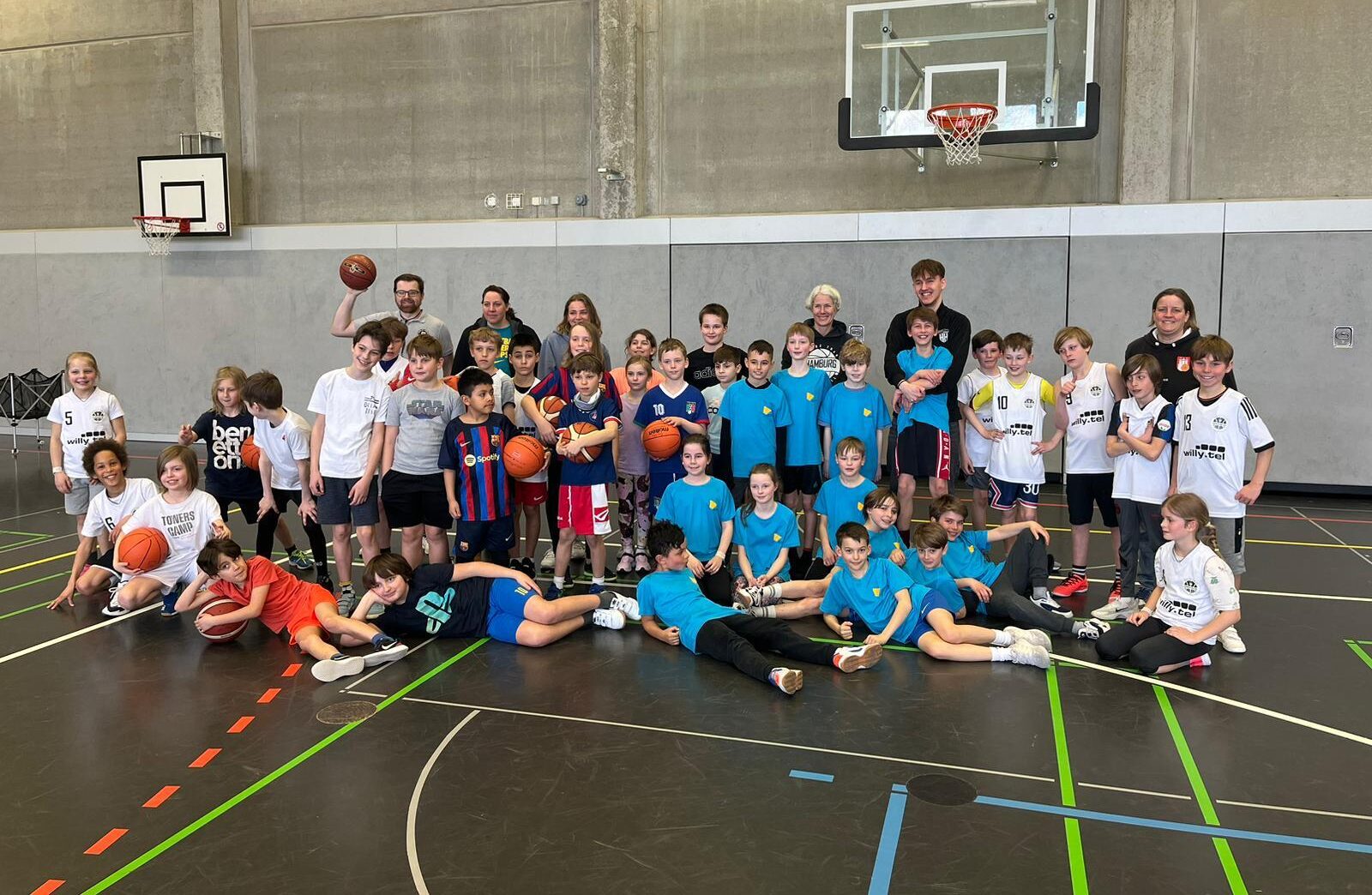 Gruppenbild von Kindern und Jugendlichen in einer Sporthalle, einige tragen blaue Trikots, andere weiße, mit Basketballkörben im Hintergrund