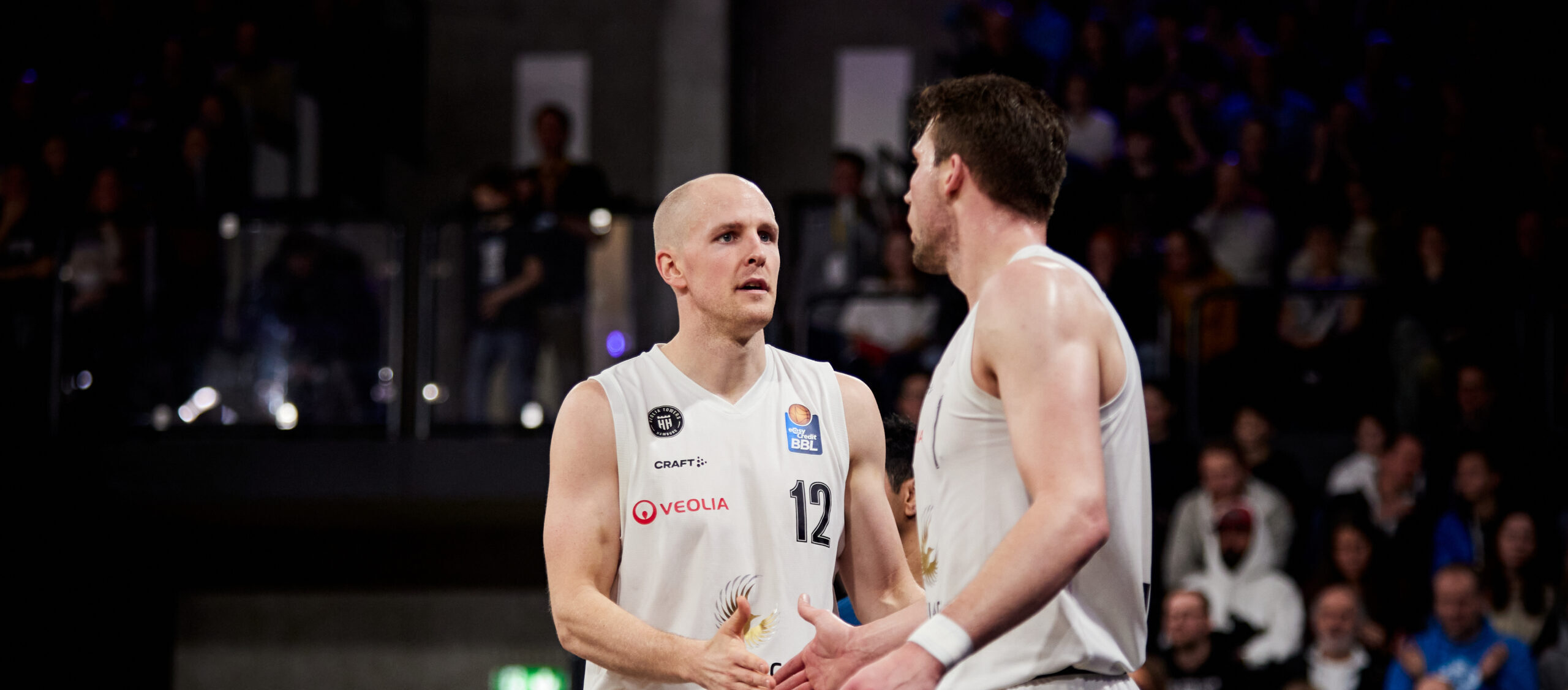 Zwei Basketballspieler der Würzburg Baskets in weißen Trikots mit den Nummern 12 und 5 während eines Spiels