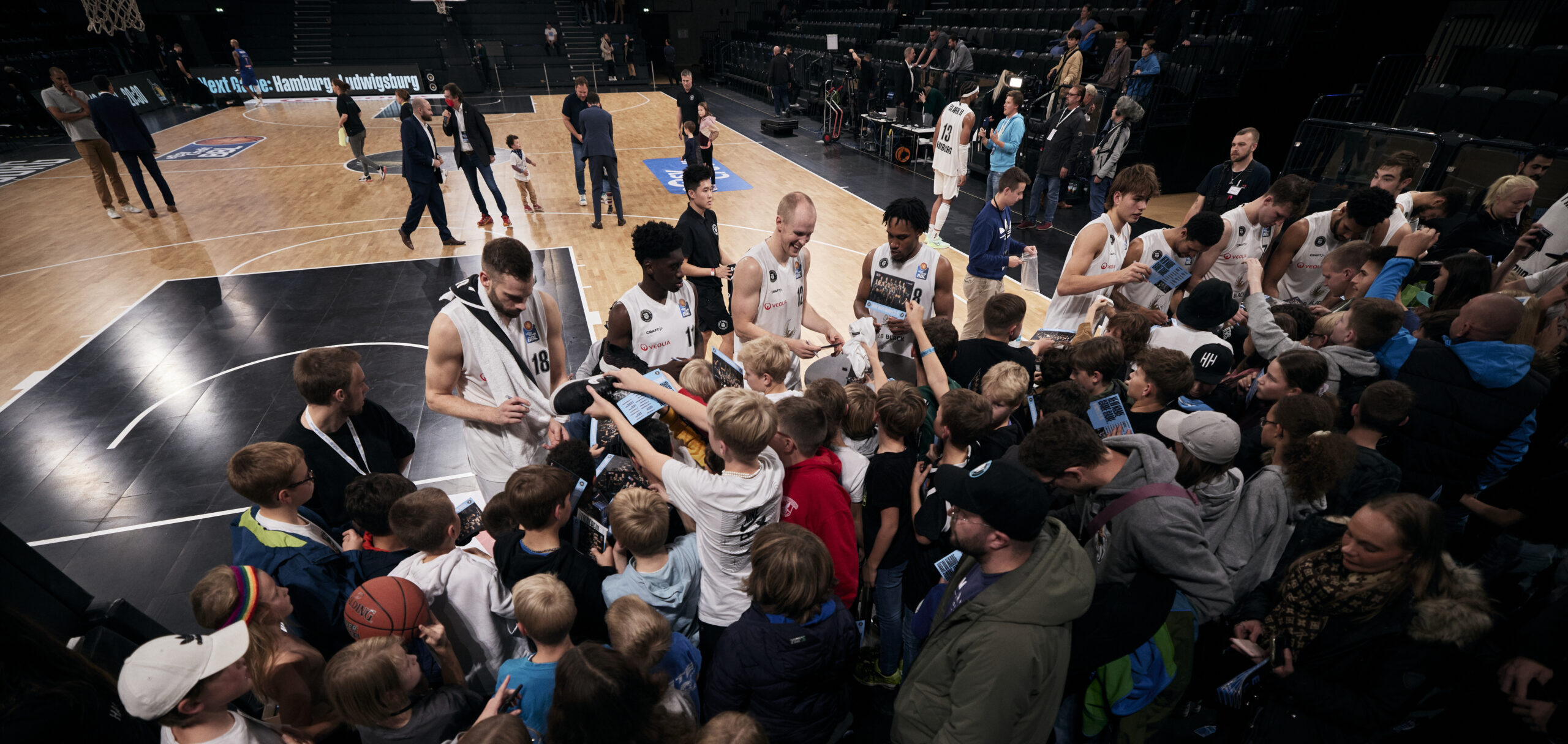 Basketballspieler in weißen Trikots geben Autogramme an eine Gruppe von Kindern und Jugendlichen am Spielfeldrand einer Halle