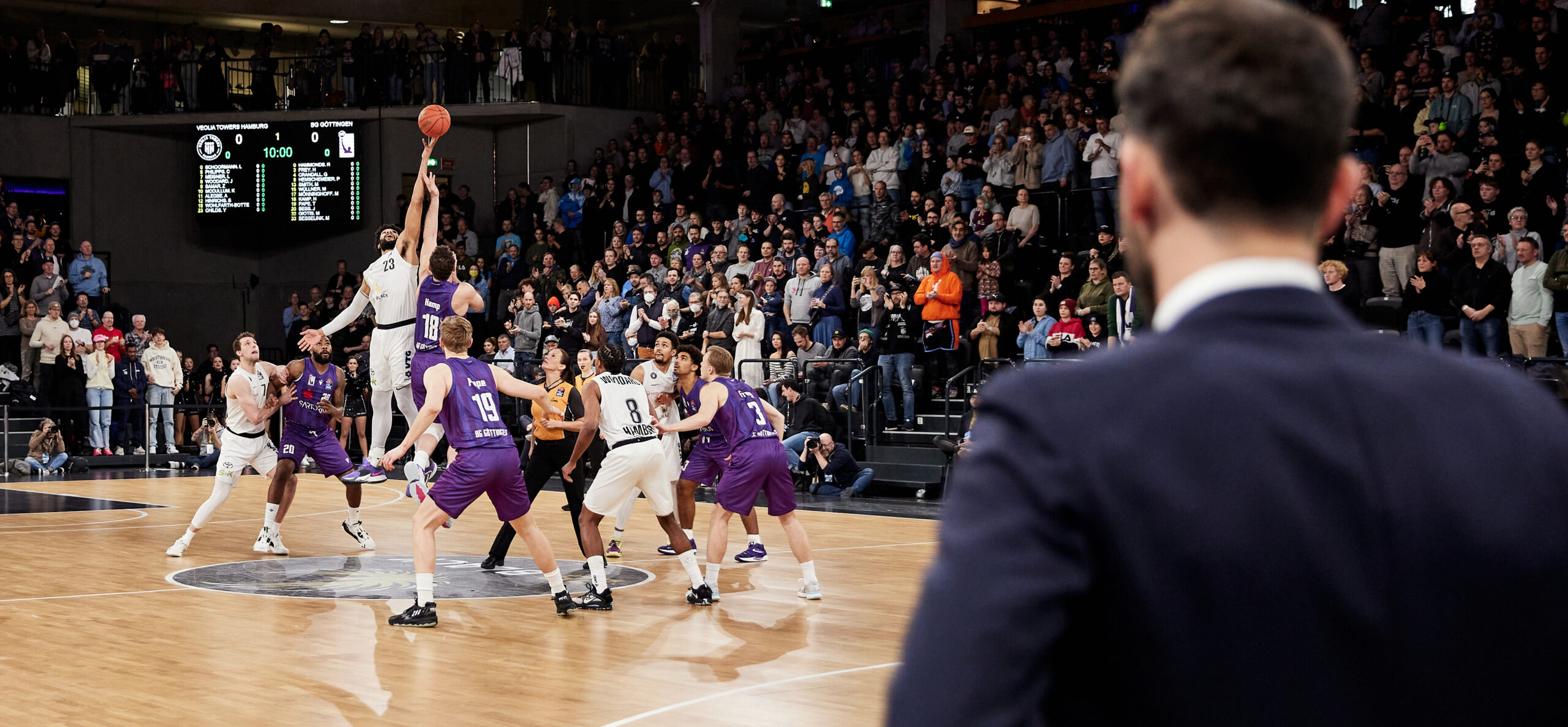 Zwei Basketballspieler springen zum Sprungball in der Mitte des Spielfelds, umgeben von weiteren Spielern und Zuschauern auf den Tribünen. Im Vordergrund, verschwommener Umriss von Mann in blauem Anzug von Hinten