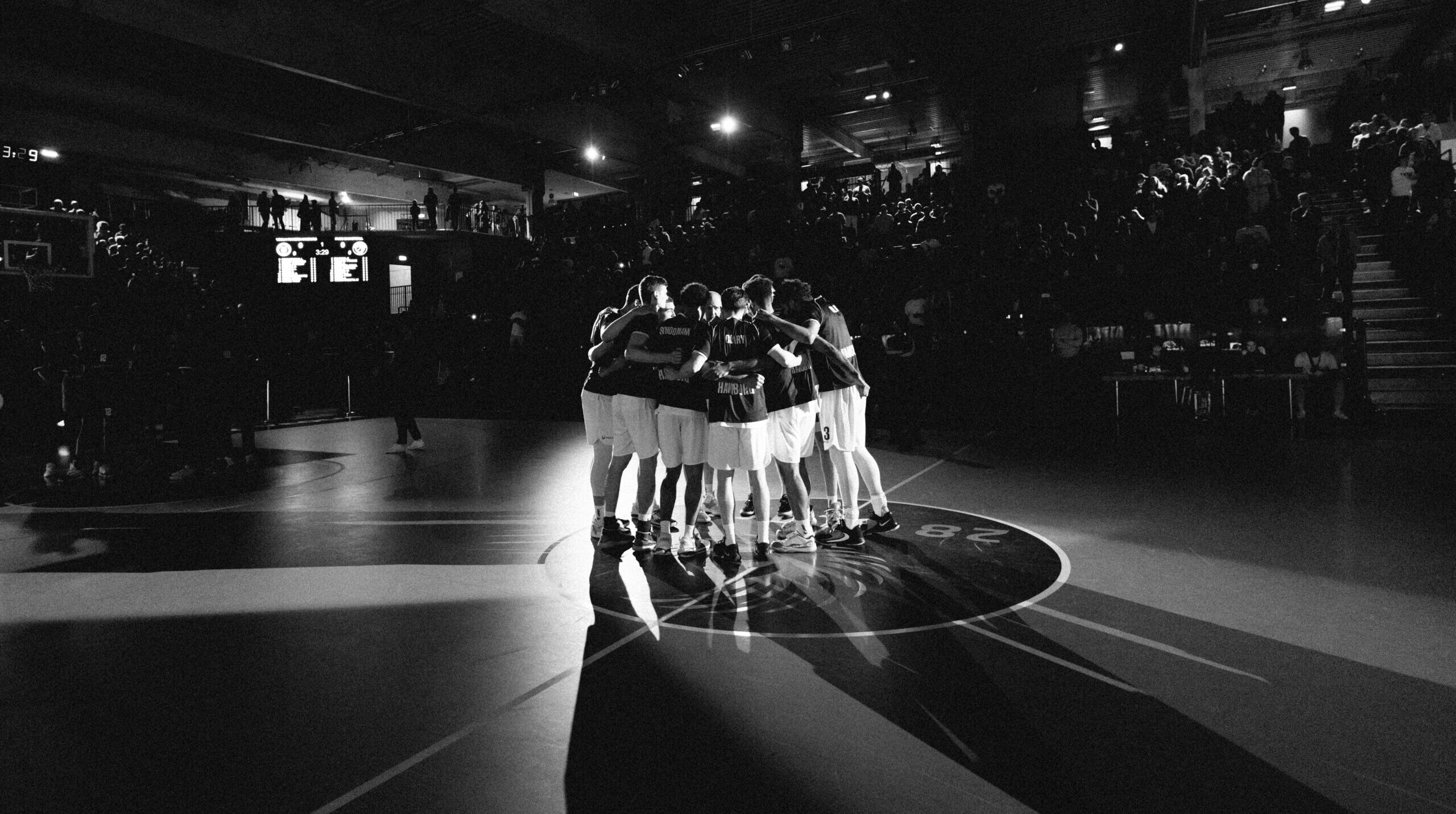 Basketballteam in weißen Shorts und dunklen Trikots steht im Kreis auf dem Spielfeld, umarmt sich vor Spielbeginn