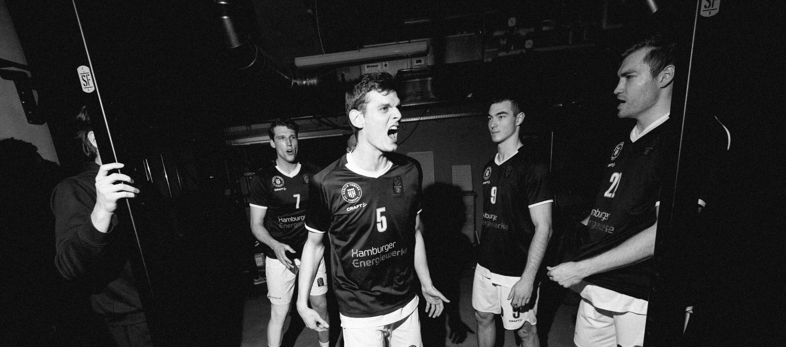 Fünf Basketballspieler der Hamburg Towers in dunklen Trikots und weißen Shorts stehen in einem dunklen Raum, bereit für das Spiel gegen Trento.