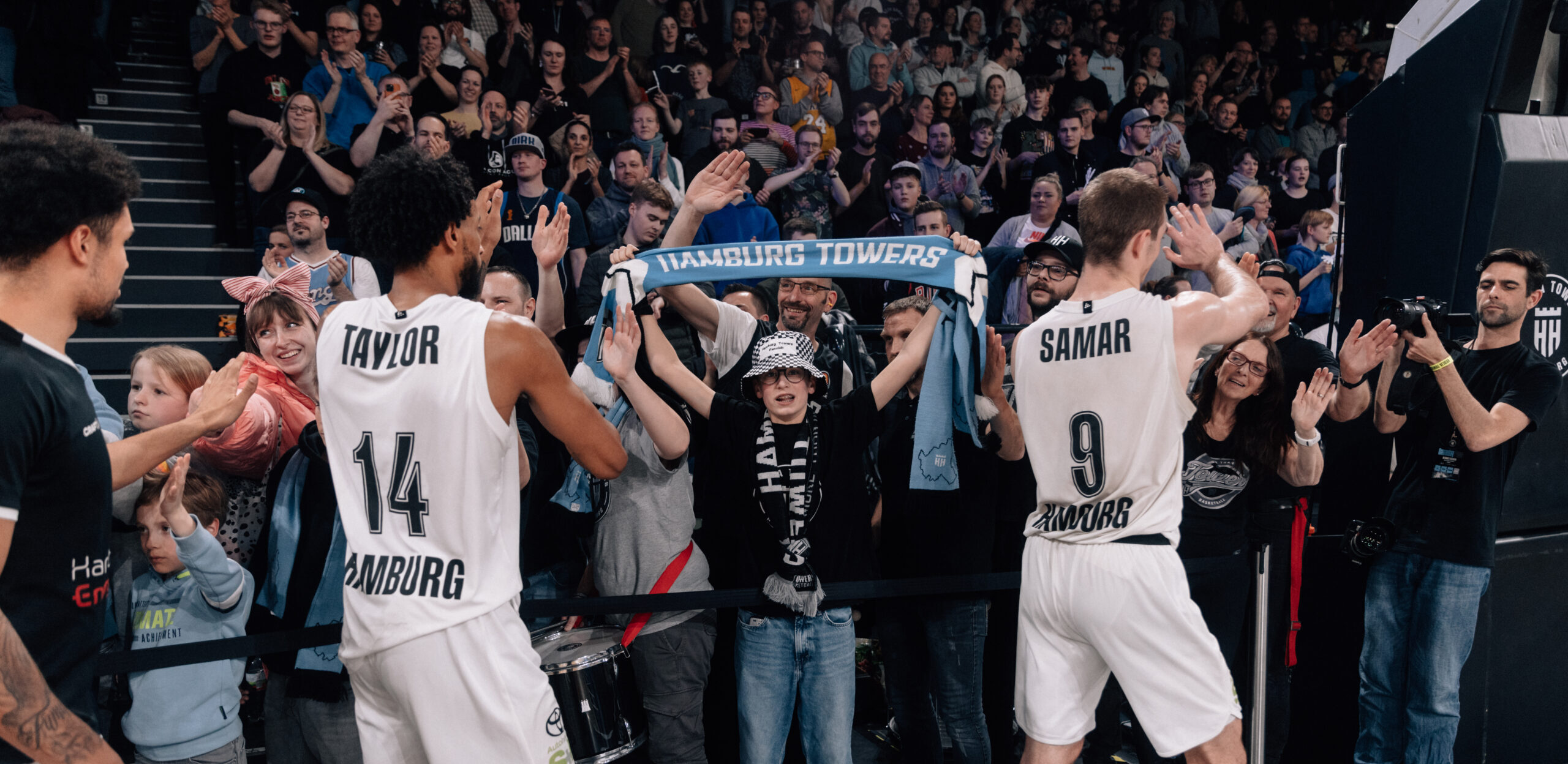 Zwei Basketballspieler der Hamburg Towers mit den Trikots Nummer 14 und 9 feiern vor jubelnden Fans in der Halle