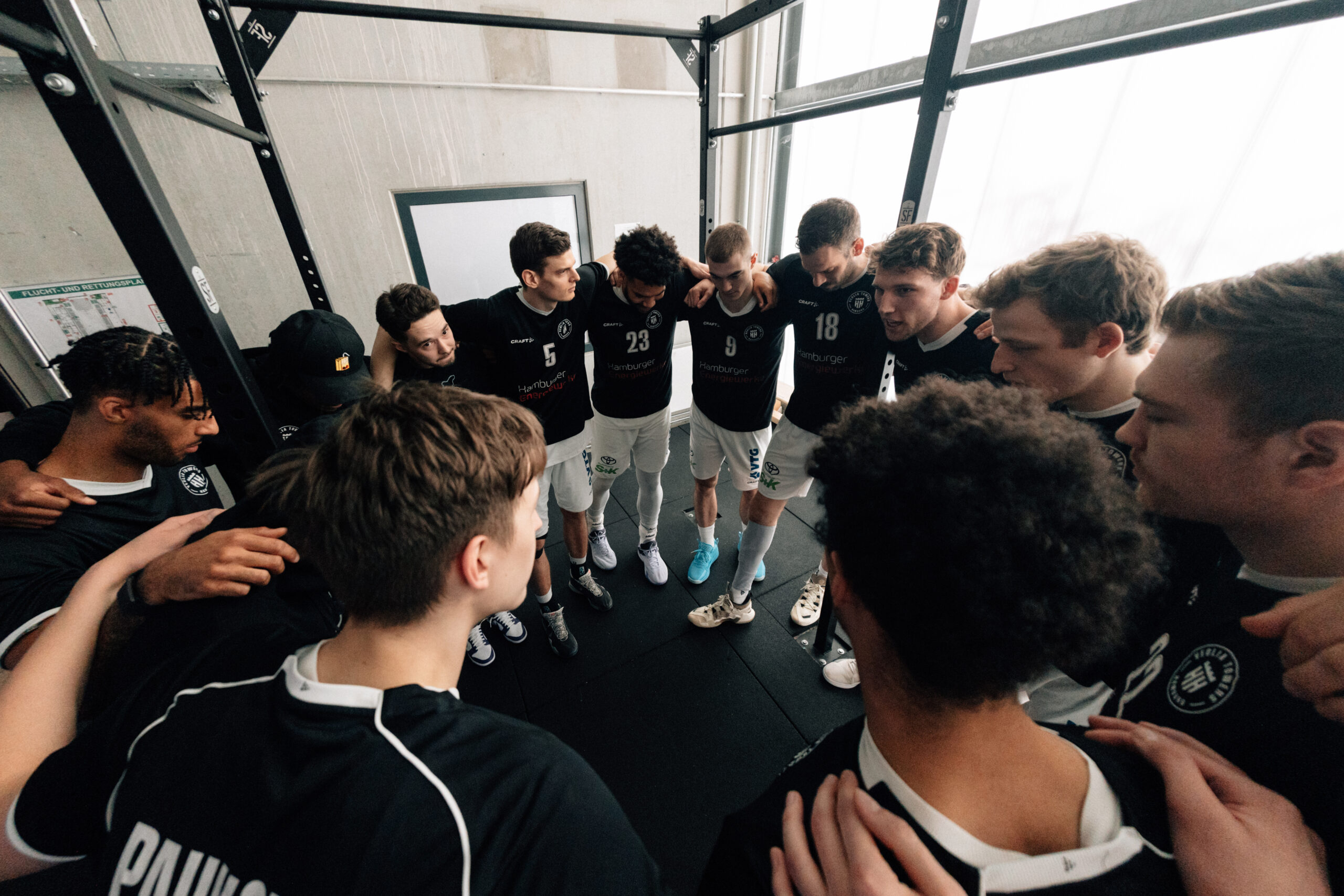 Basketballteam in weißen Shorts und dunklen Trikots steht im Kreis, umarmt sich vor Spielbeginn