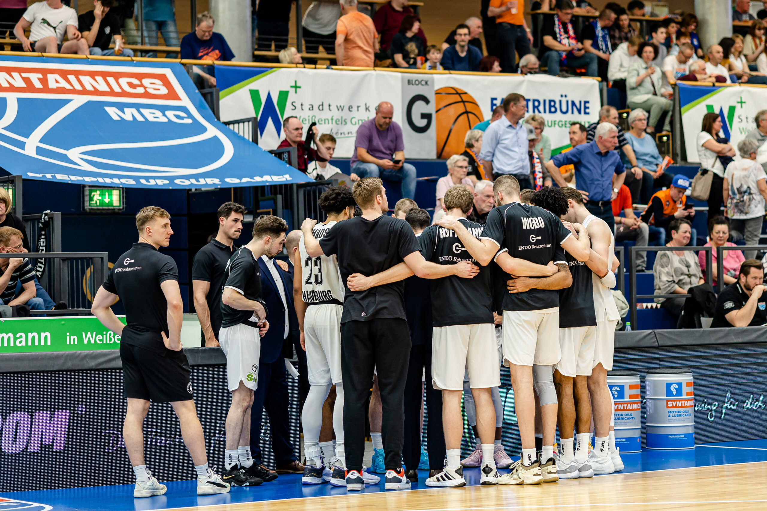 Basketballteam in weißen Shorts und dunklen Trikots steht im Kreis auf dem Spielfeld, umarmt sich vor Spielbeginn
