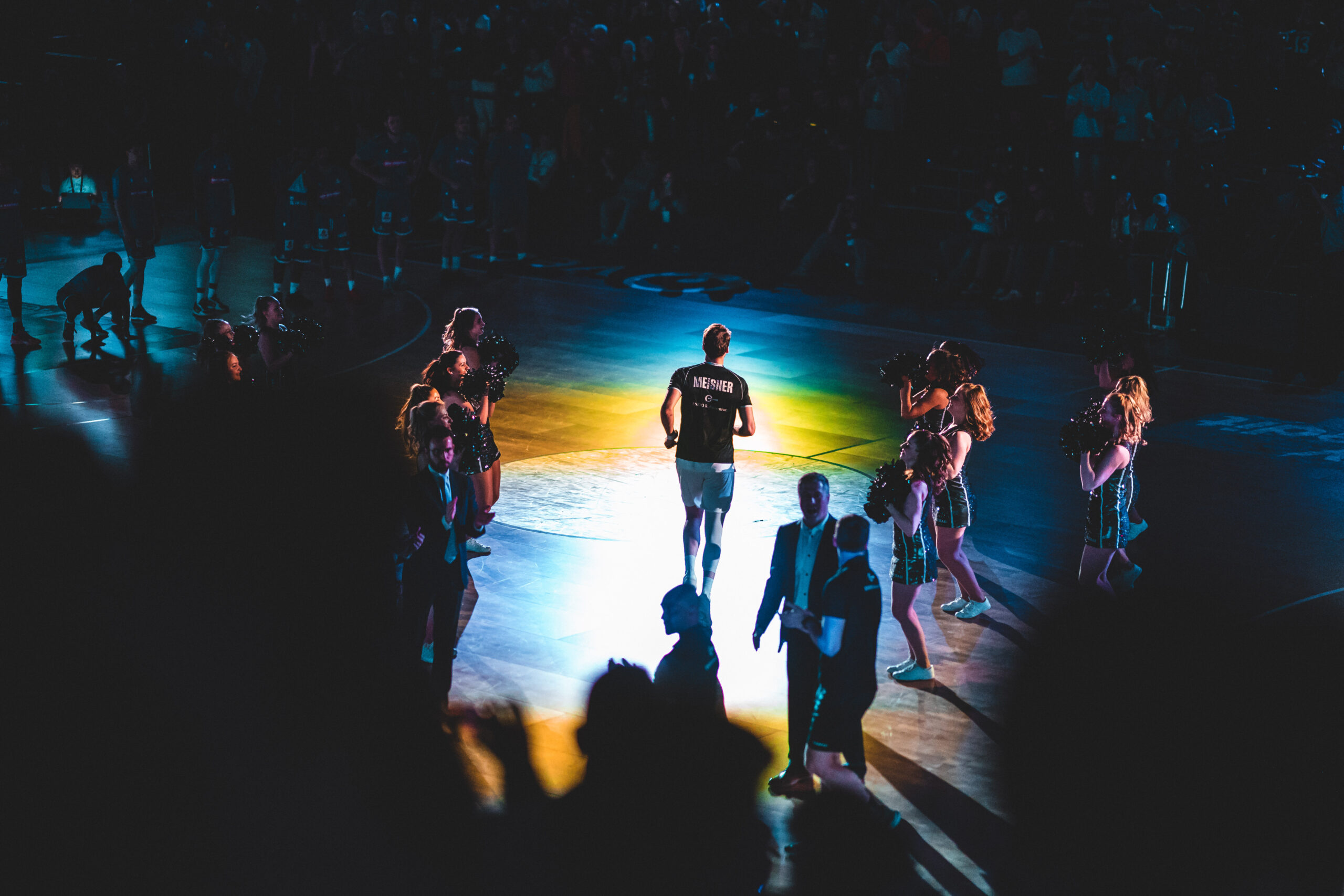 Basketballspieler läuft auf Spielfeld, umgeben von Cheerleadern und Publikum im dunklen Stadionlicht