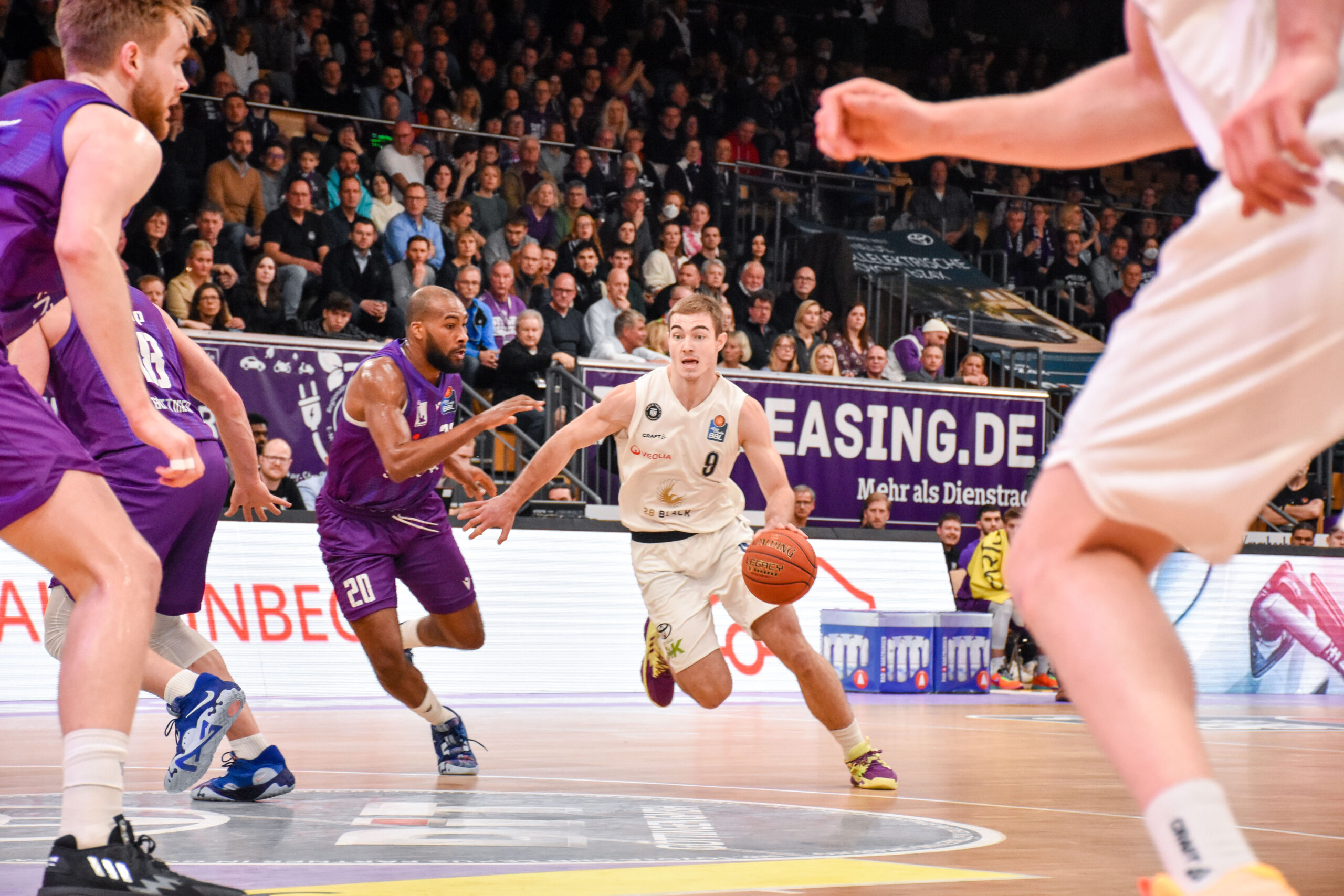 Spieler in weißem Trikot dribbelt den Ball, Spieler in lila Uniform verteidigt aktiv, Zuschauer im Hintergrund