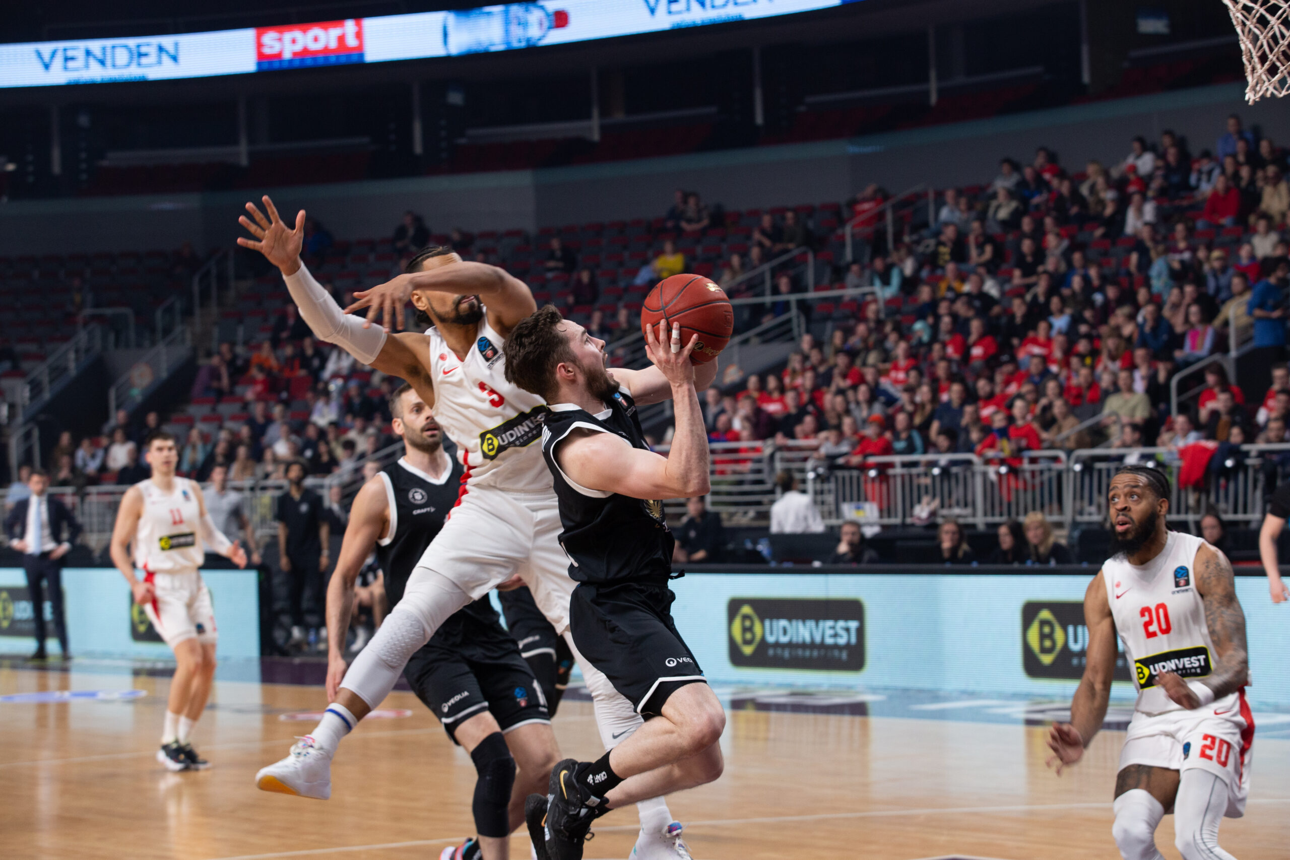 Basketballspieler in schwarzem Trikot springt zum Korb, während Spieler in weißen Trikots verteidigen, Zuschauer im Hintergrund