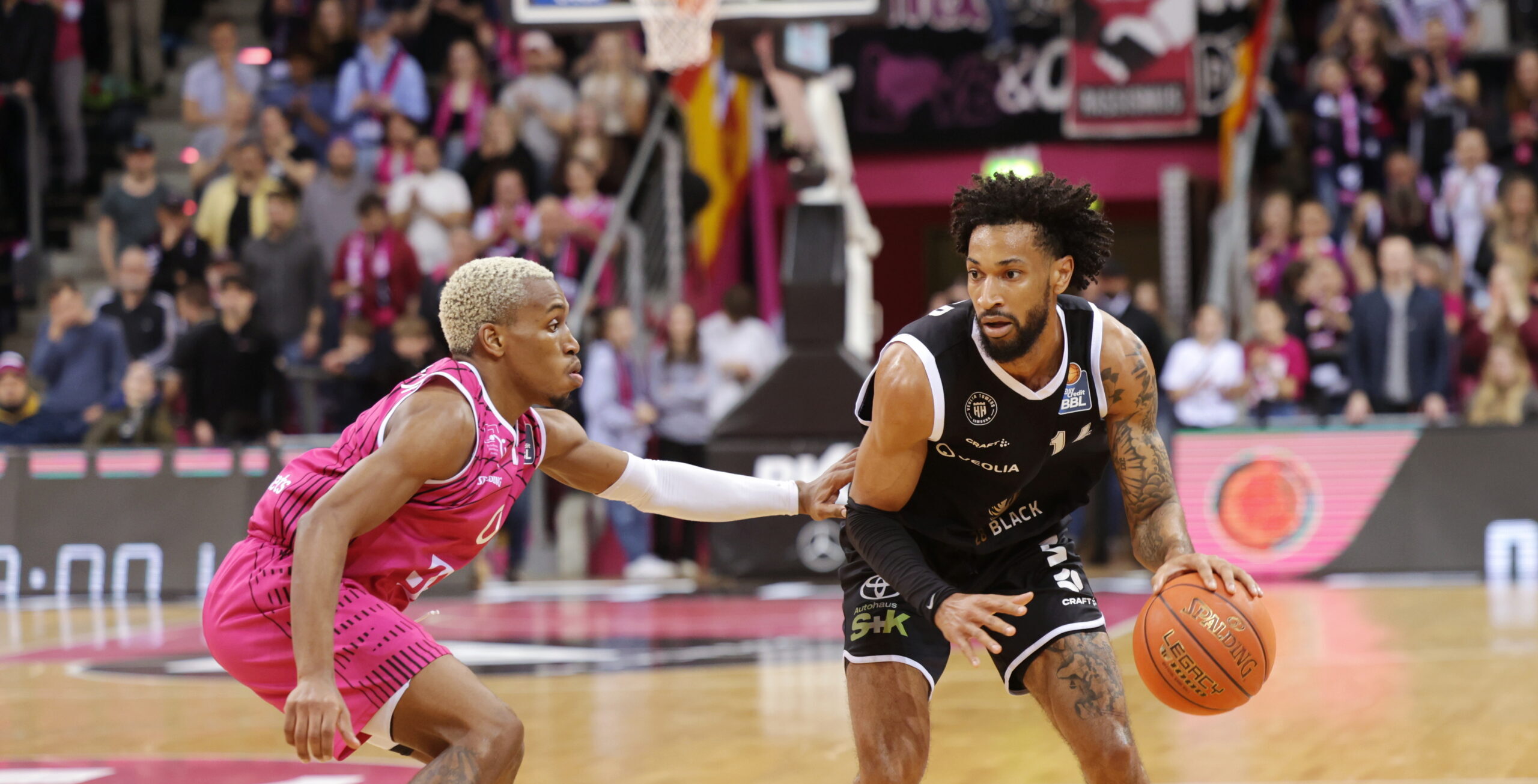 Zwei Basketballspieler im Spiel, einer in schwarz dribbelt den Ball, der andere in pink verteidigt nah, Zuschauer im Hintergrund