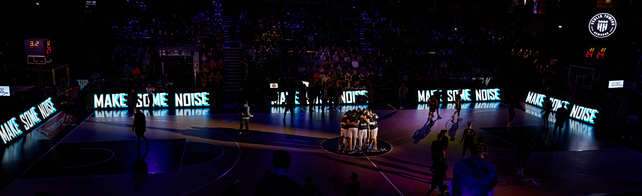 Spielfeld im Dunkeln, auf den LED-Banden steht "MAKE SOME NOISE", ein Basketballteam in der Mitte des Spielfelds in schwarzen und weißen Shirts steht eng beieinander.Im Dunkeln kann man auf der Tribüne Zuschauer erkennen