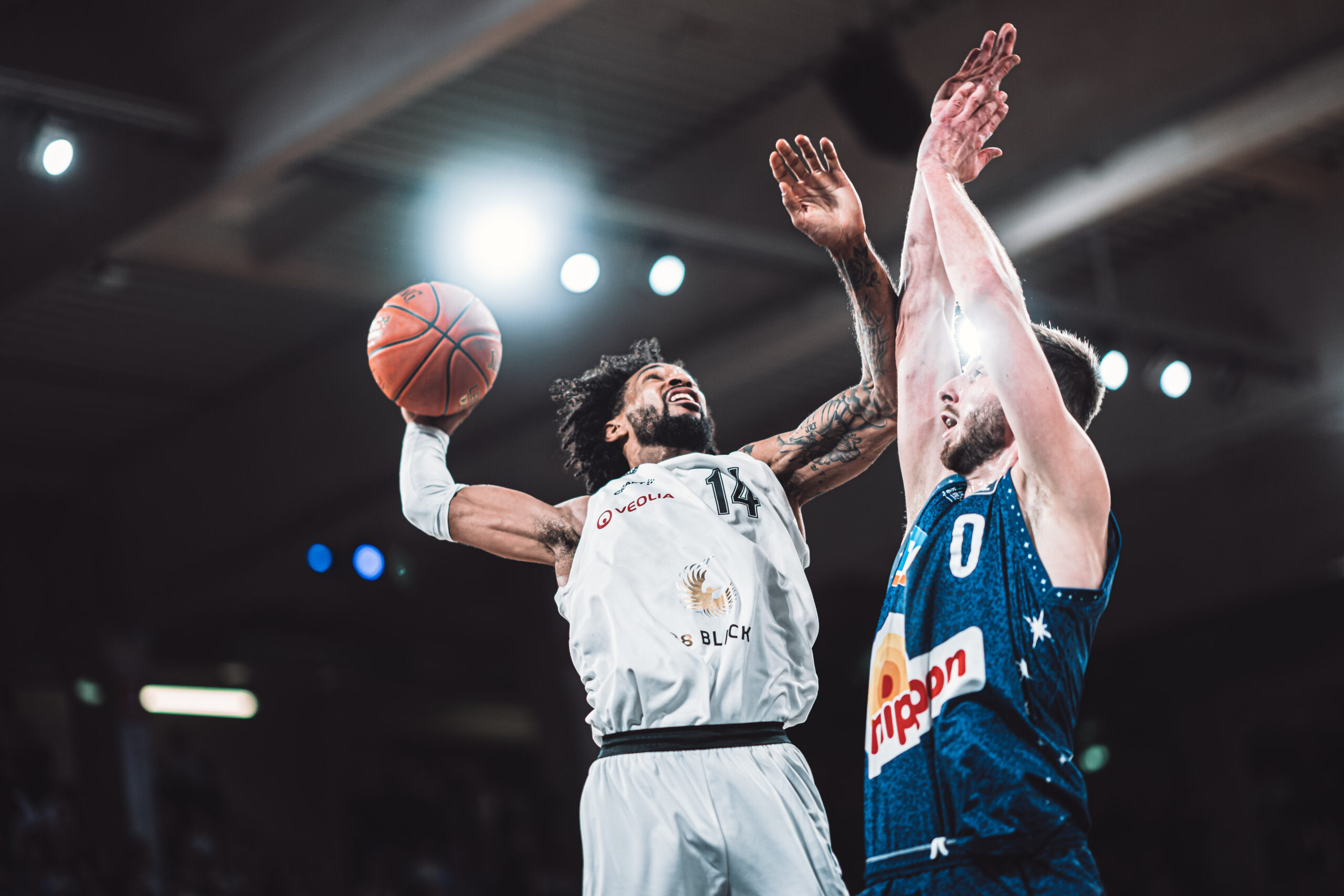 Basketballspieler in weißem Trikot springt zum Korb, verteidigt von Spieler in blauem Trikot mit erhobenen Armen