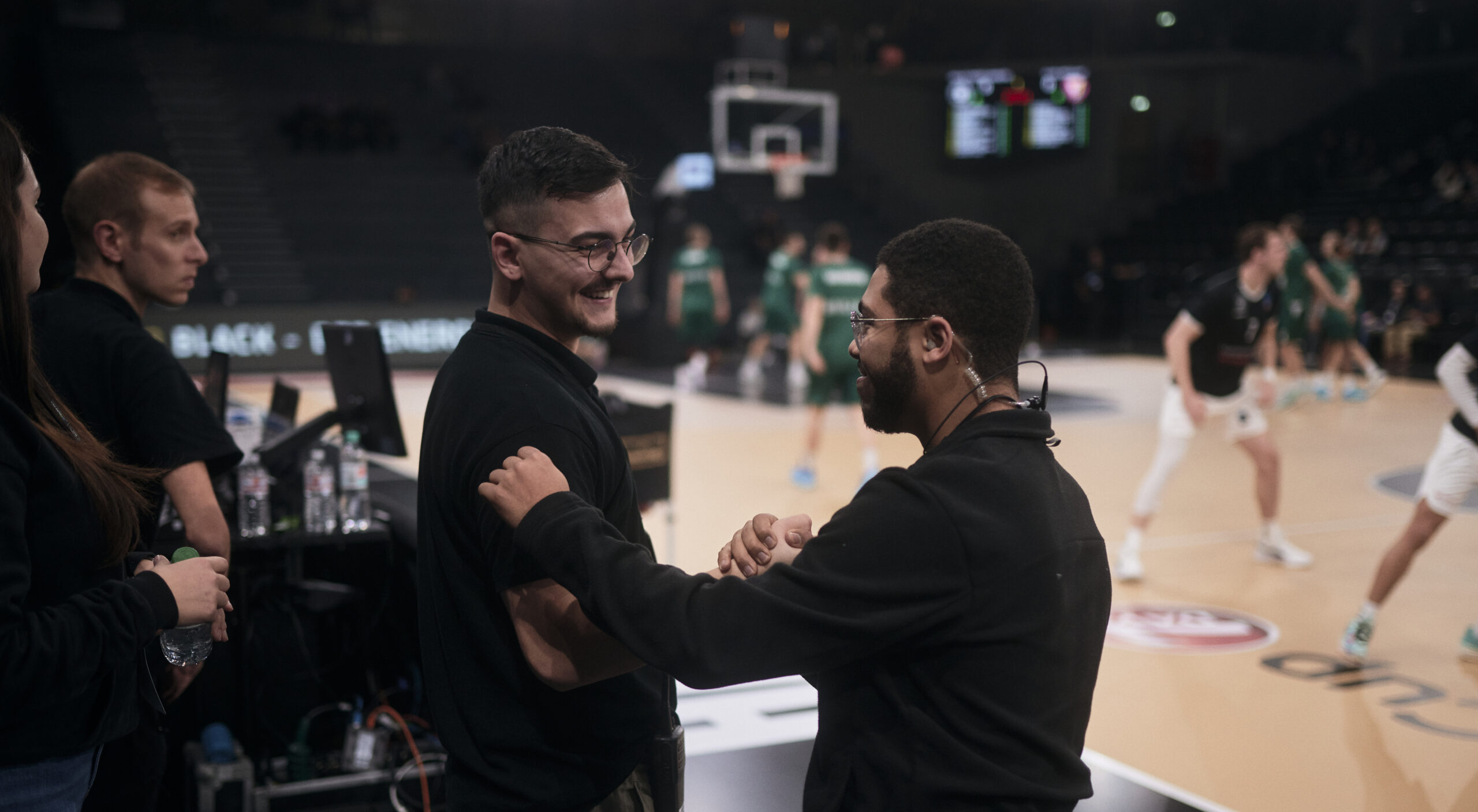 Zwei Männer in schwarzer Kleidung stehen am Spielfeldrand einer Basketballhalle, im Hintergrund spielen Spieler in grünen und weißen Trikots auf dem Basketballfeld.