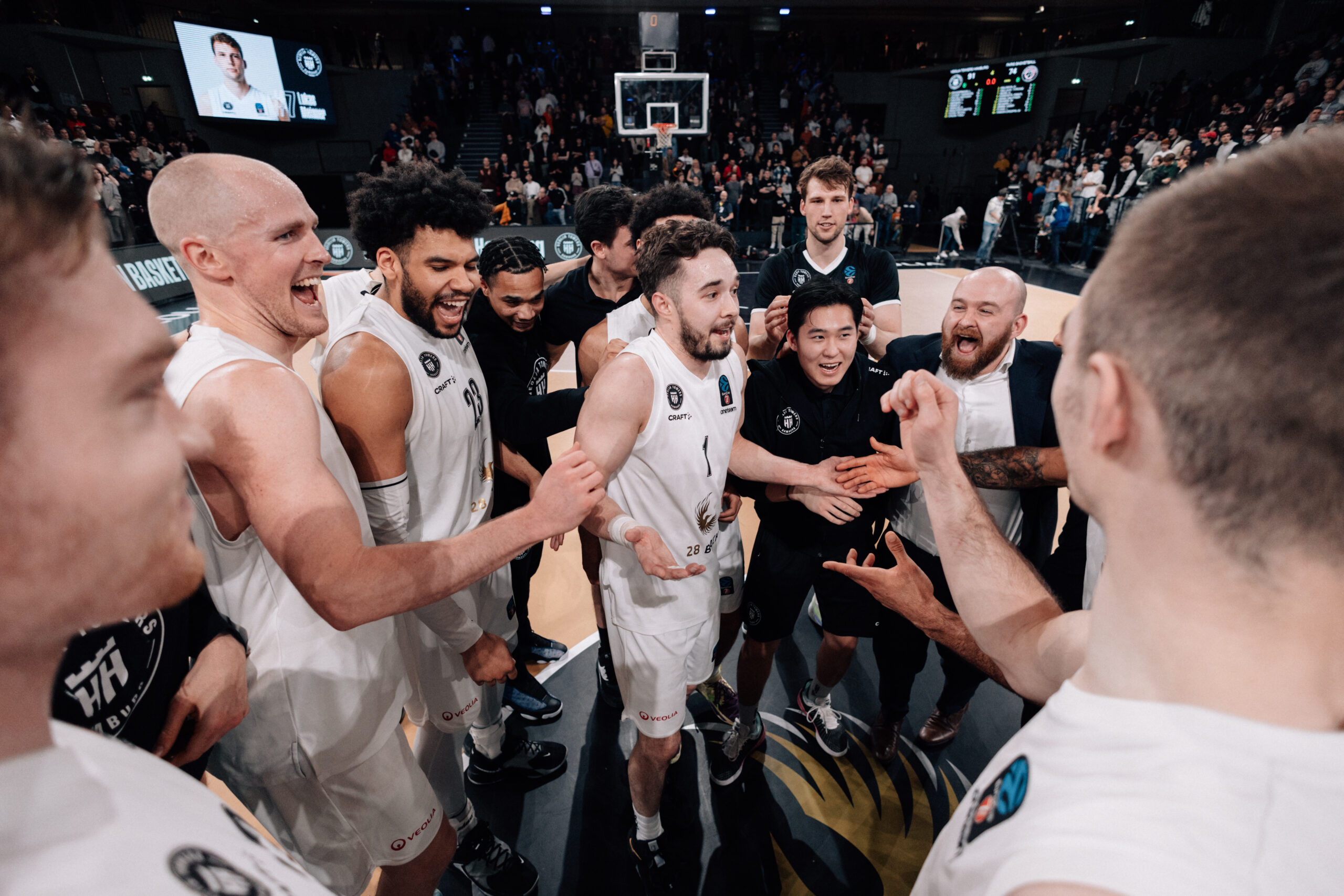 Basketballspieler der Towers in weißen Trikots versammeln sich in einer Arena während eines Spiels gegen Paris, umgeben von Zuschauern.