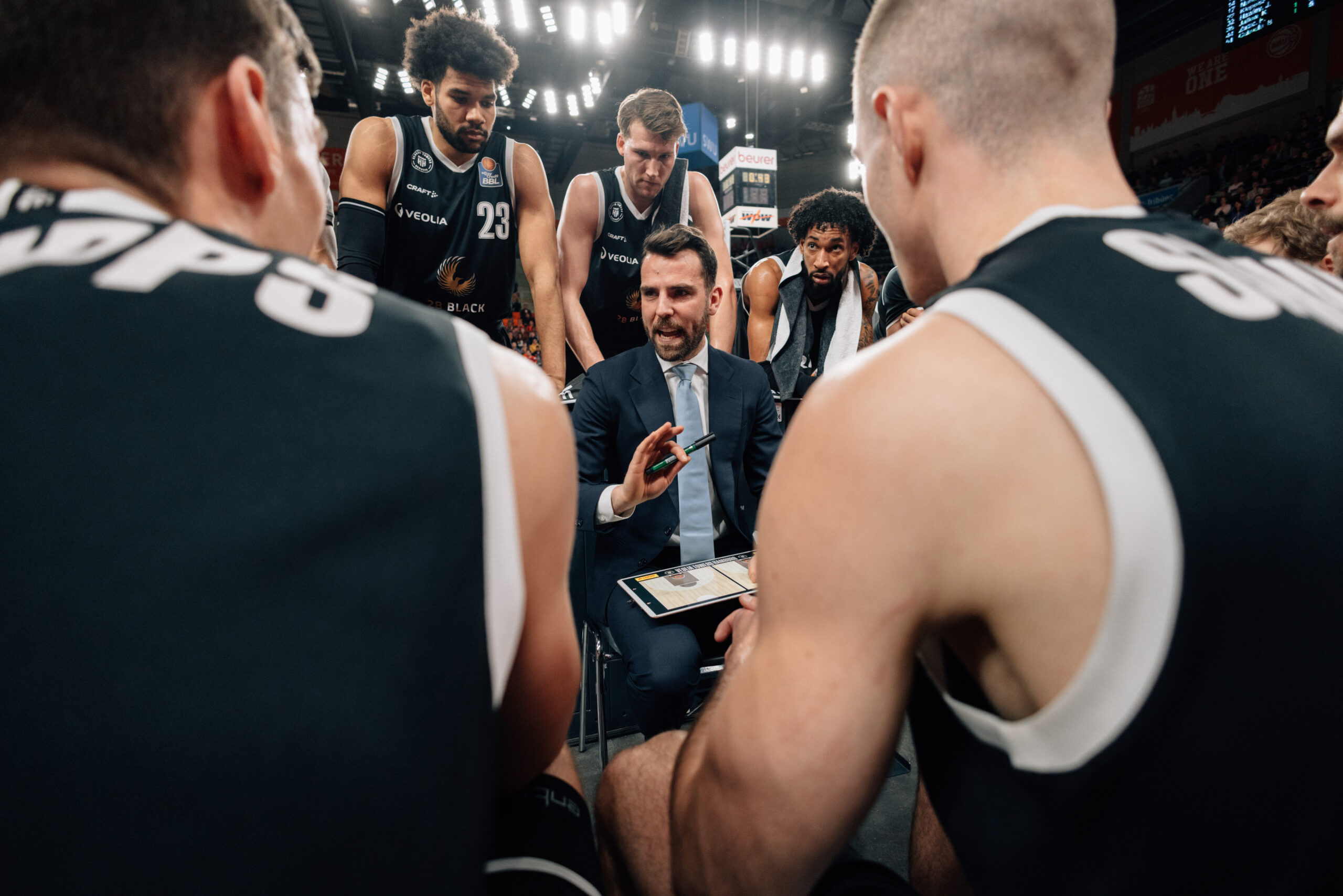 Basketballtrainer im Anzug gibt Anweisungen während einer Auszeit, umgeben von Spielern in schwarzen Trikots