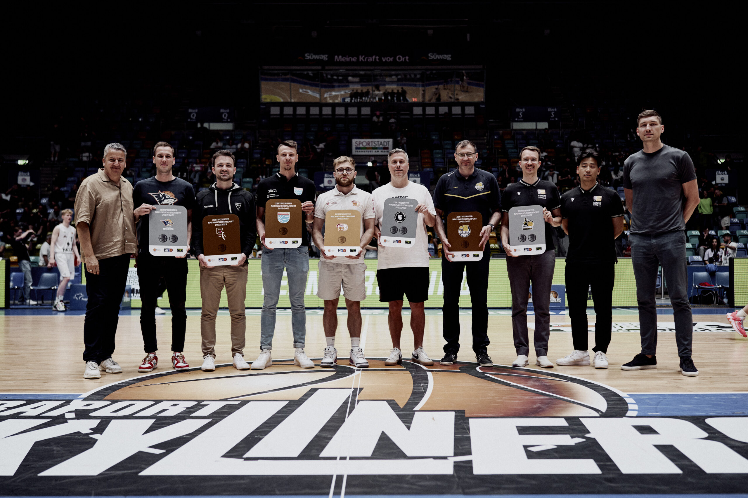 Zehn Männer stehen in einer Reihe auf einem Basketballfeld mit dem Schriftzug 'Sparr X Liners', jeder hält eine Trophäe oder Auszeichnung in der Hand.