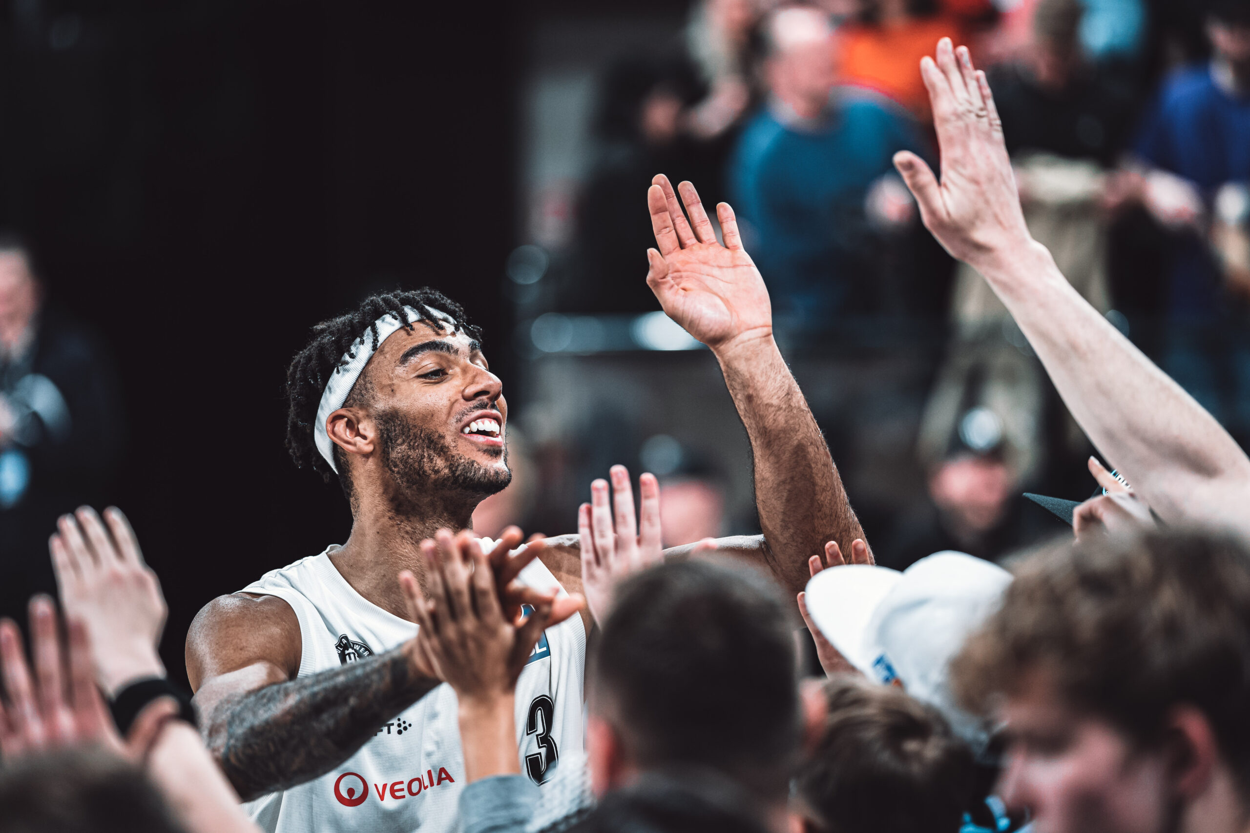 Basketballspieler in weißem Trikot mit erhobenen Händen, umgeben von Teamkollegen und Fans bei einem Spiel