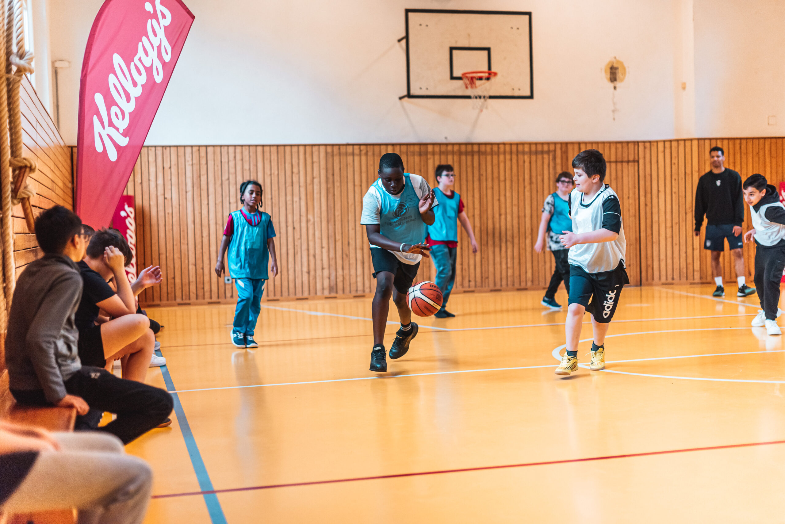 Kinder spielen Basketball in einer Halle, ein Junge dribbelt den Ball, andere beobachten und spielen mit