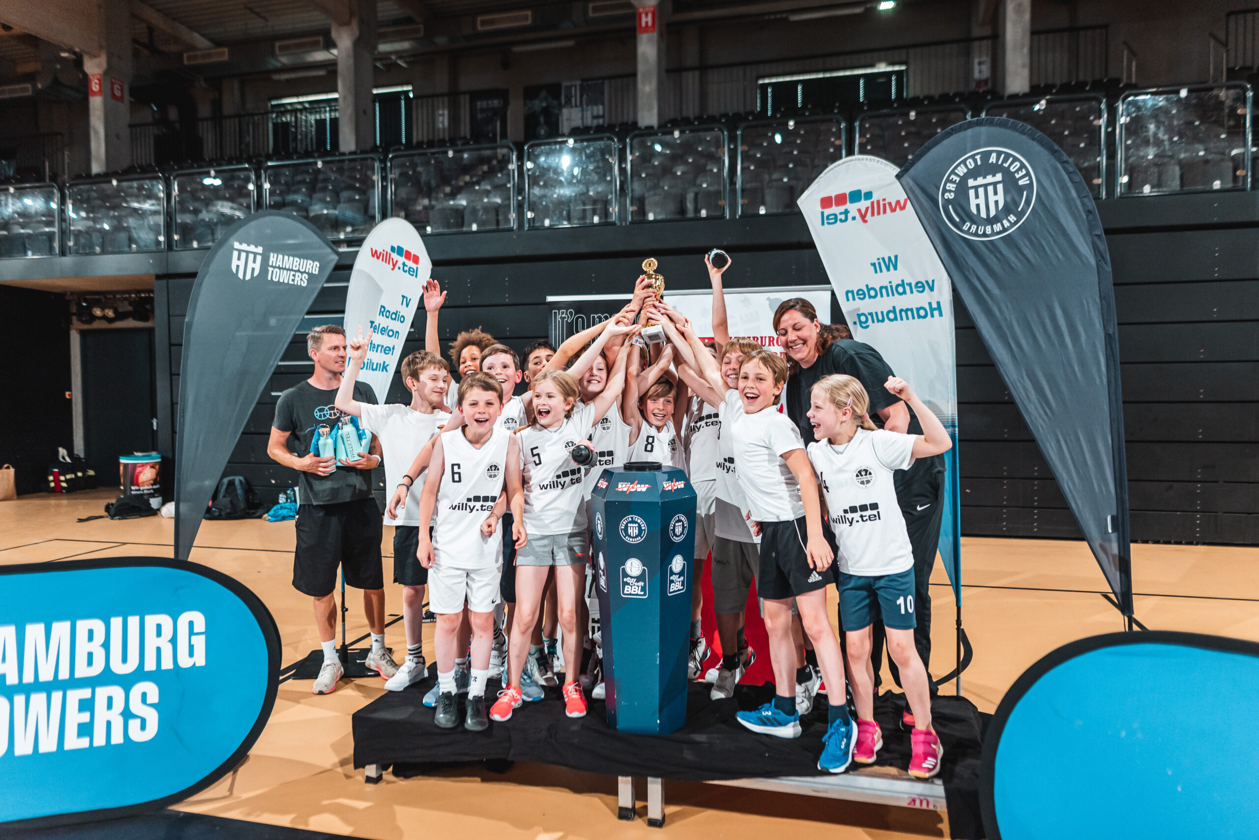 Kinder in weißen Trikots jubeln und halten gemeinsam einen Pokal in die Höhe. Im Hintergrund Banner von den Hamburg Towers und Willy.tel