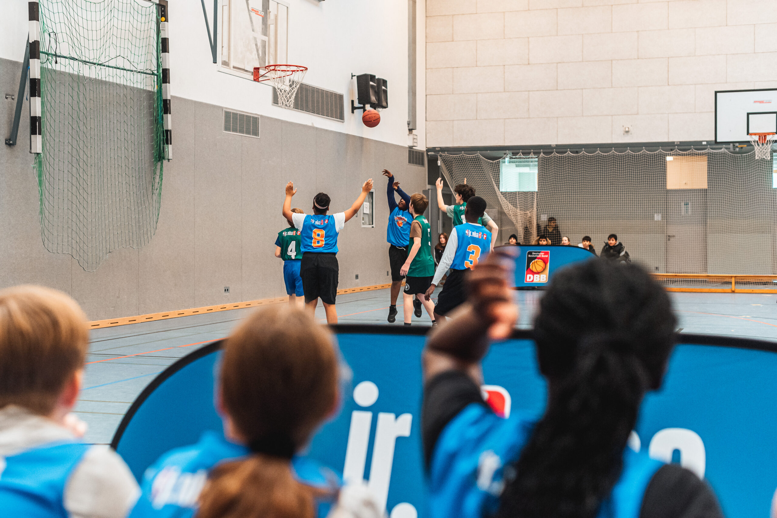 Jugendliche spielen Basketball in einer Halle, ein Spieler wirft den Ball zum Korb, andere versuchen zu blocken