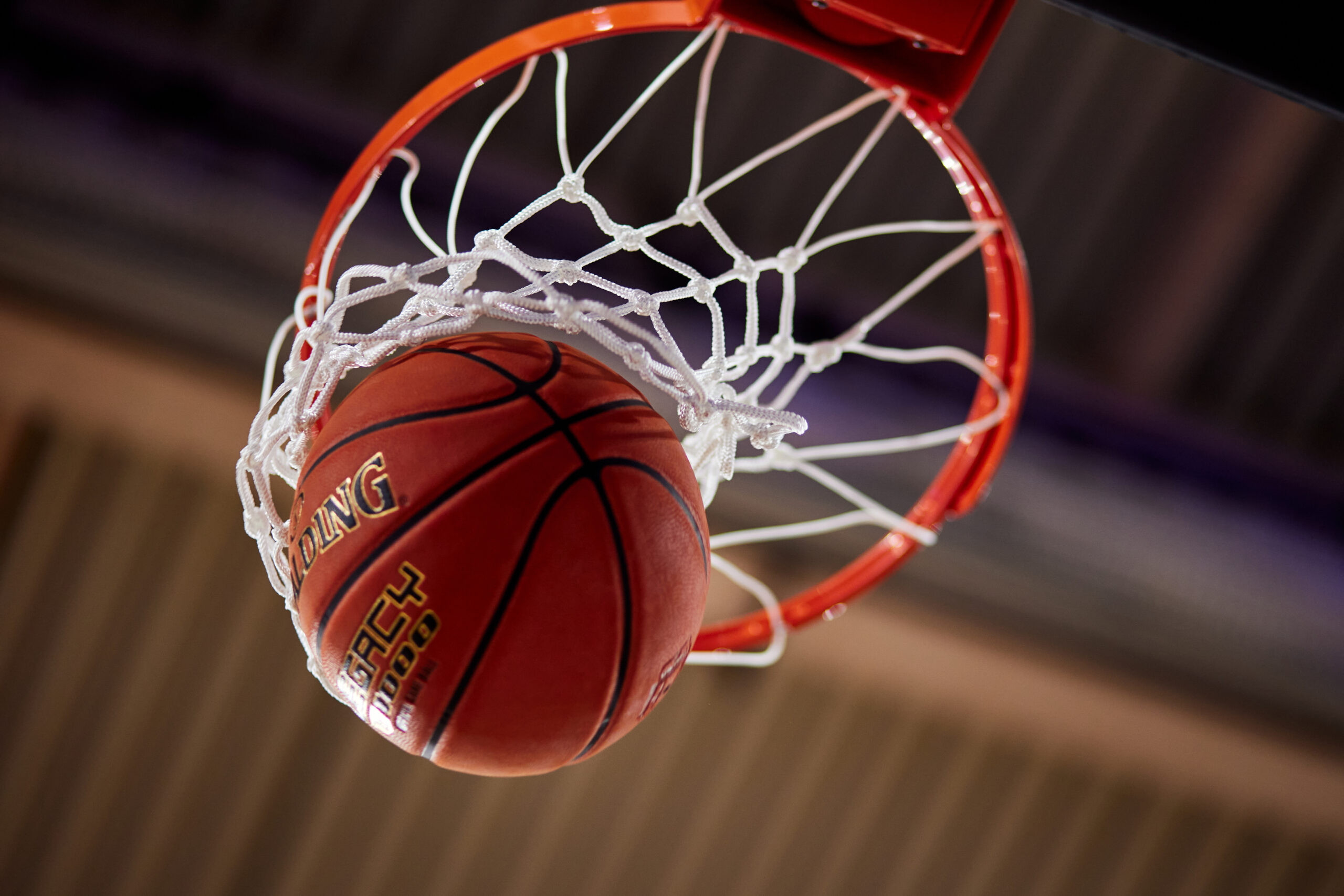 Ein orangener Spalding Basketball fliegt durch den Ring und das Netz eines Basketballkorbs, im Hintergrund eine Hallendecke